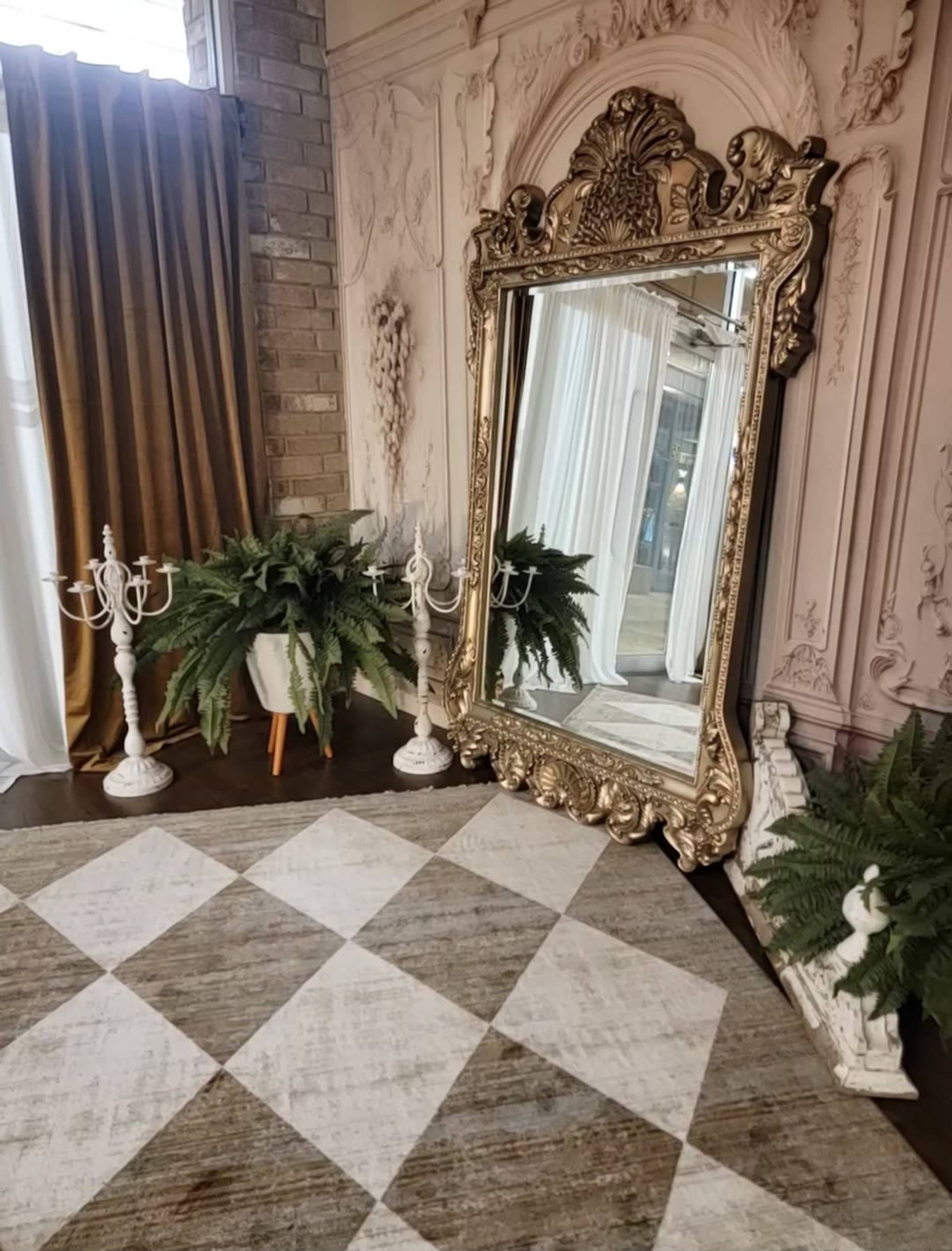 The scene features a large ornate mirror next to potted ferns and candelabra on a checkered rug in a beautifully decorated room with tall curtains.