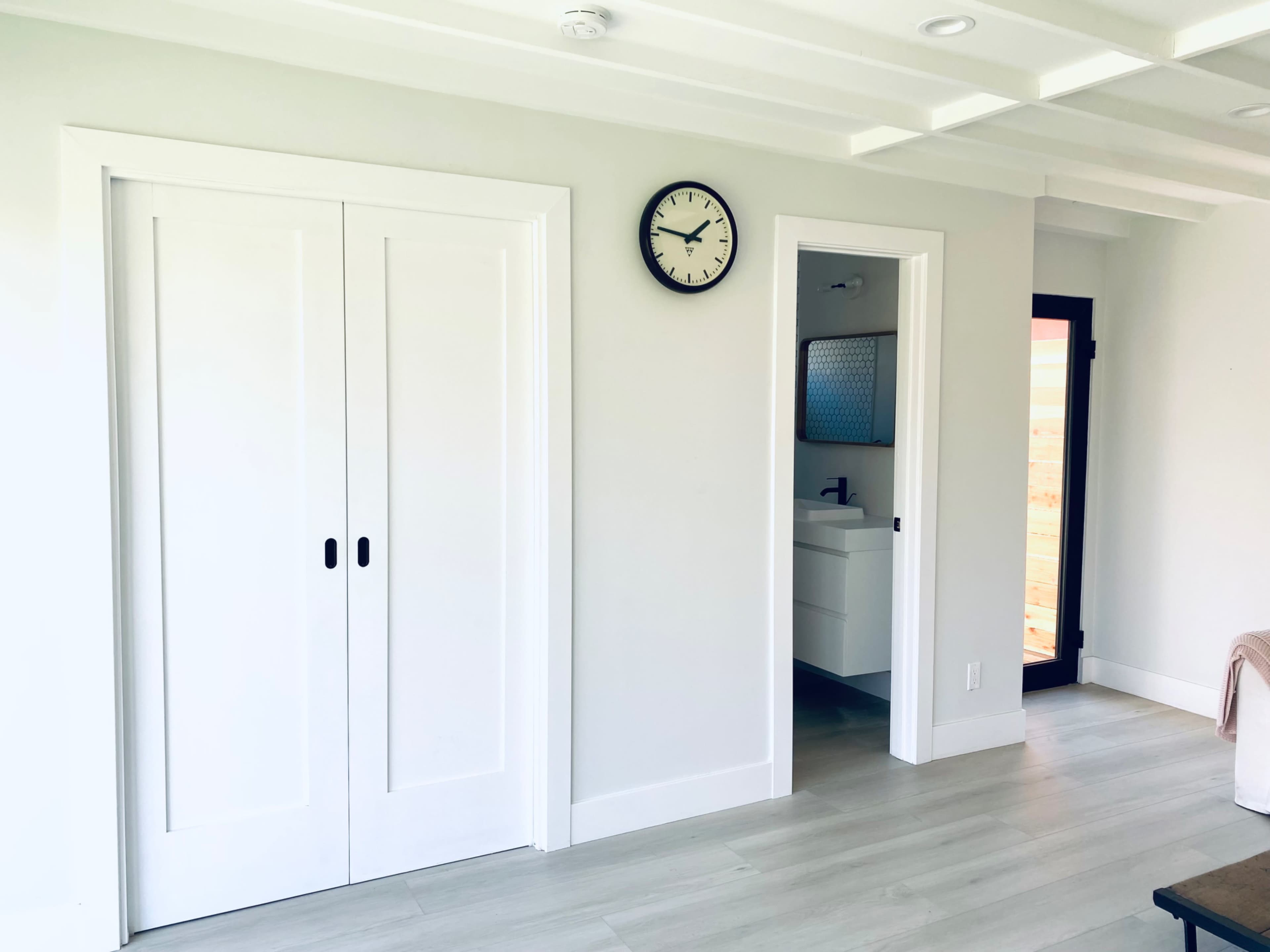 The image shows a light-colored room with a wall clock and two doorways, one leading to a bathroom and the other with sliding doors.
