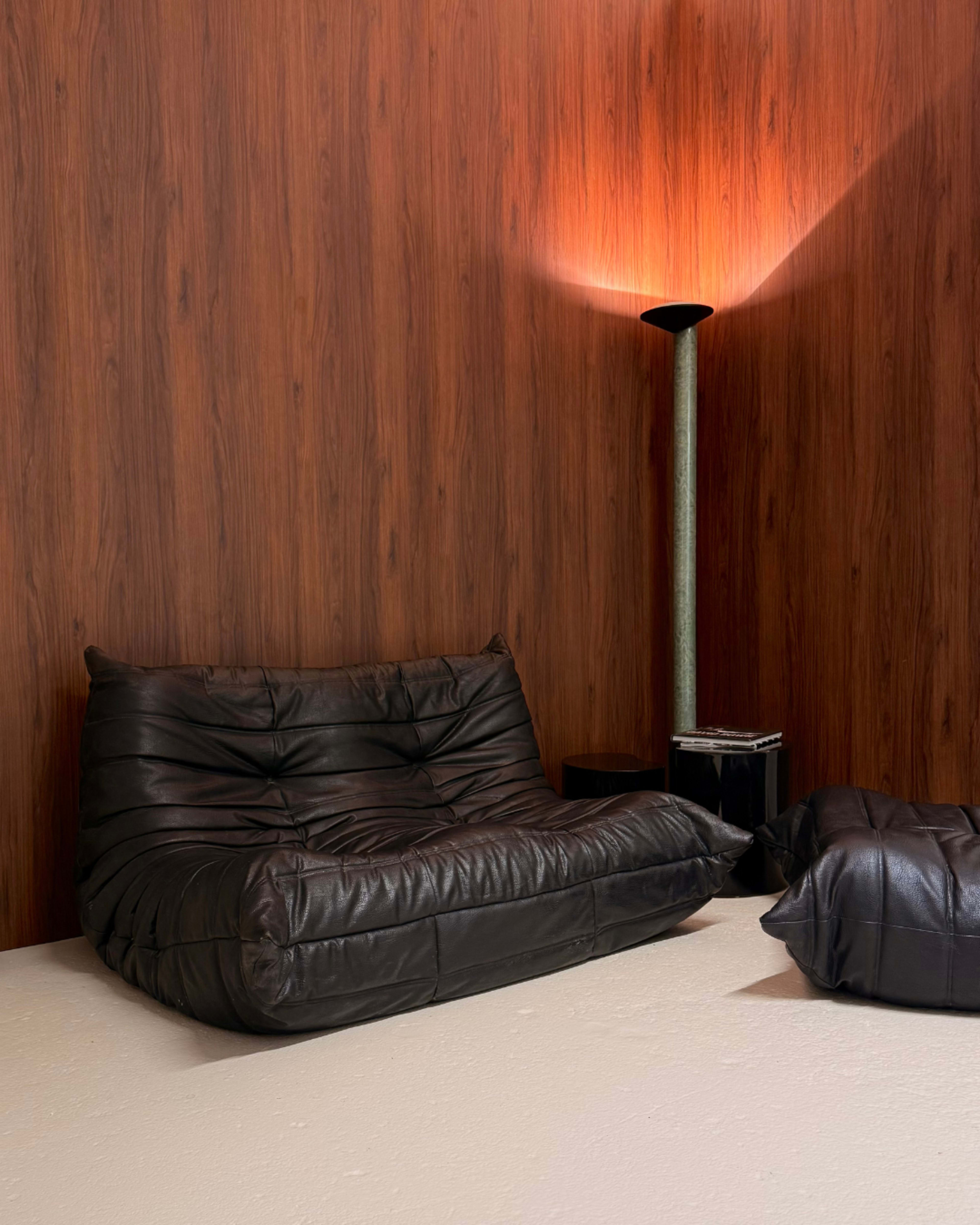 The image shows a dark leather bean bag chair and a smaller matching pouf, set against a textured wooden wall, with a tall lamp providing ambient lighting.