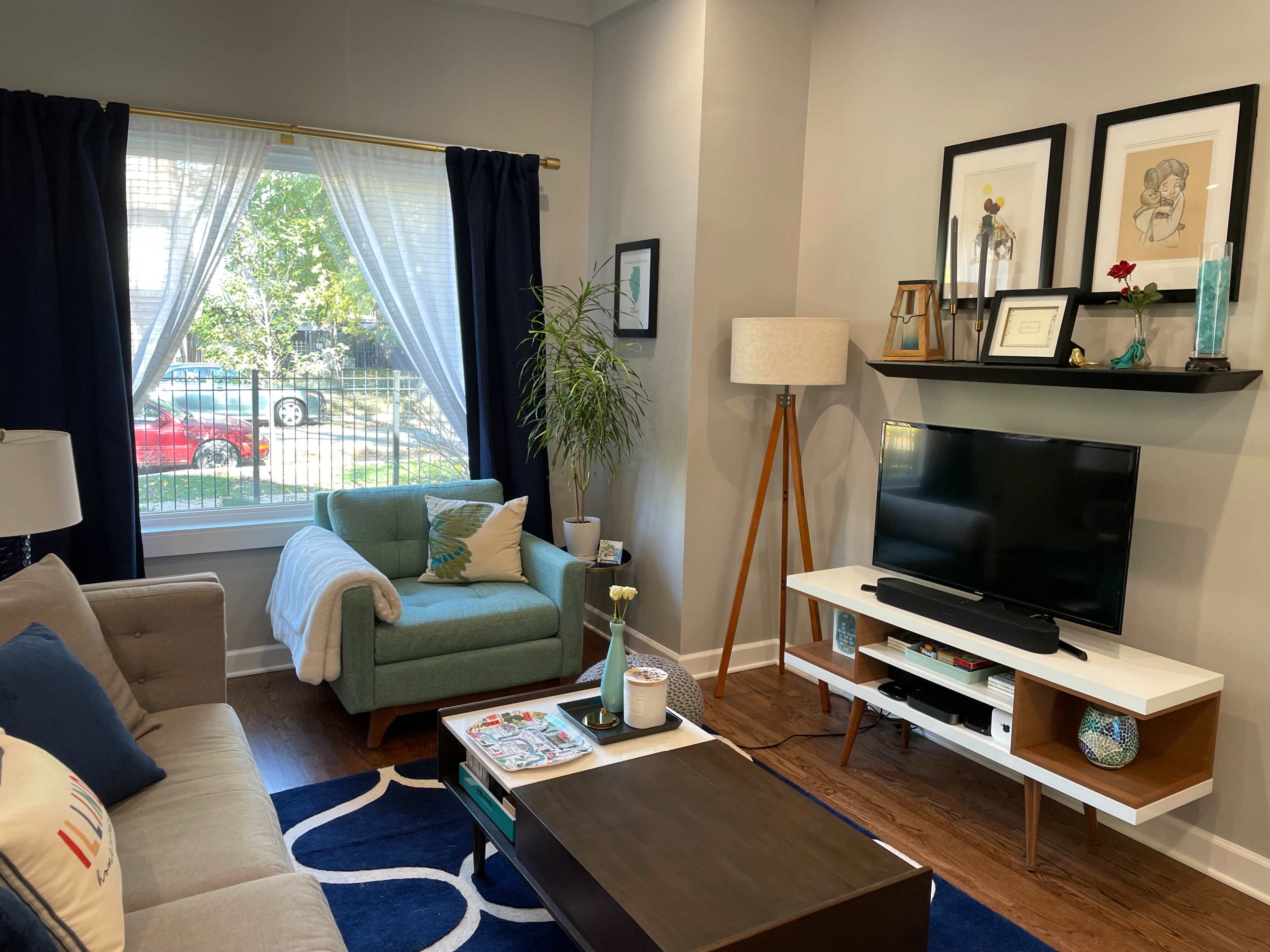 The living room features a gray sofa, a light blue armchair, a television on a white media console, and large windows with sheer curtains.