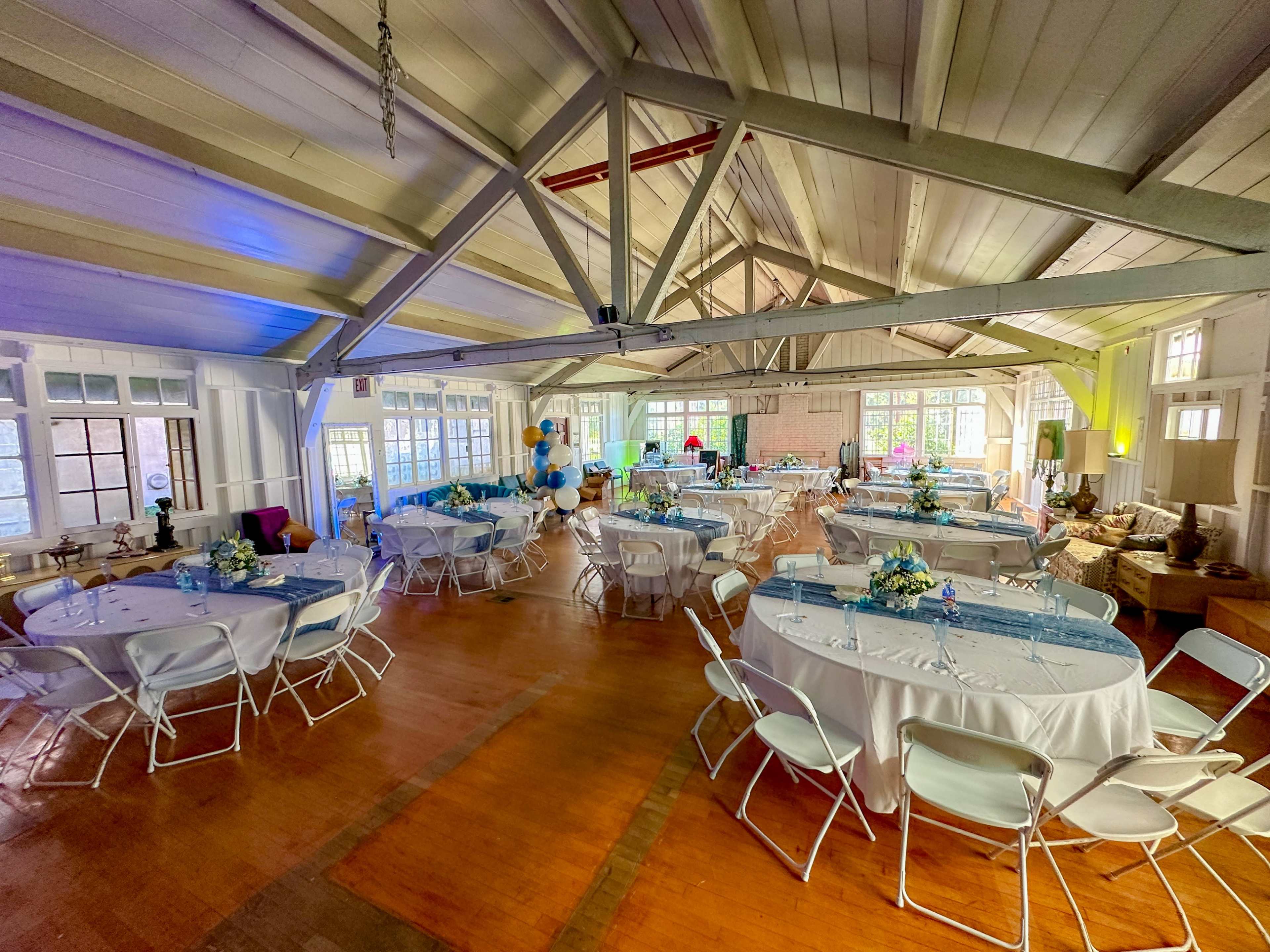 The image shows a spacious indoor venue set up for an event, featuring round tables with white tablecloths, blue decorations, and a wooden floor beneath a beamed ceiling.