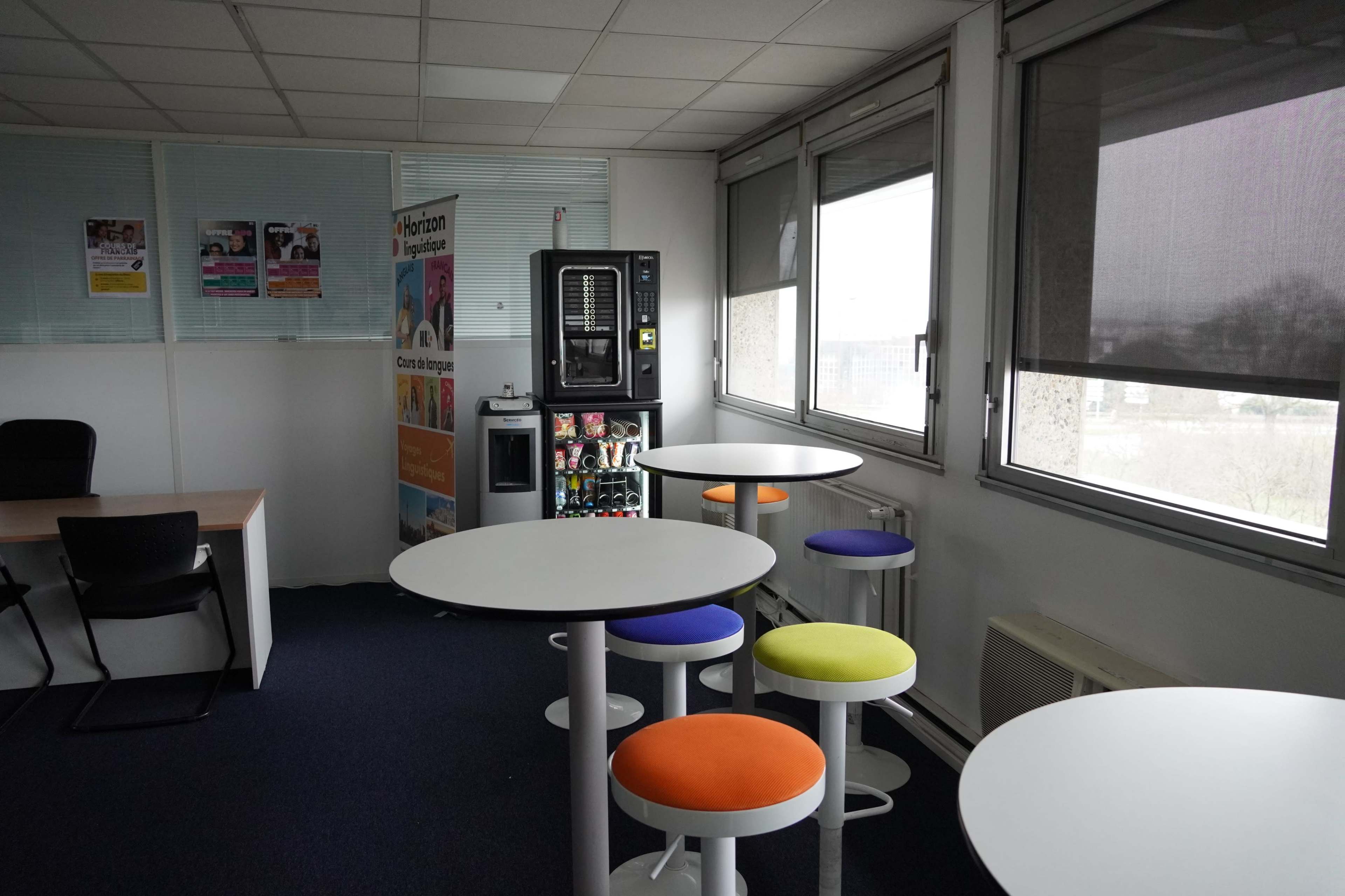 The image shows a spacious office break area featuring modern round tables with colorful stools, a vending machine, and large windows allowing natural light.