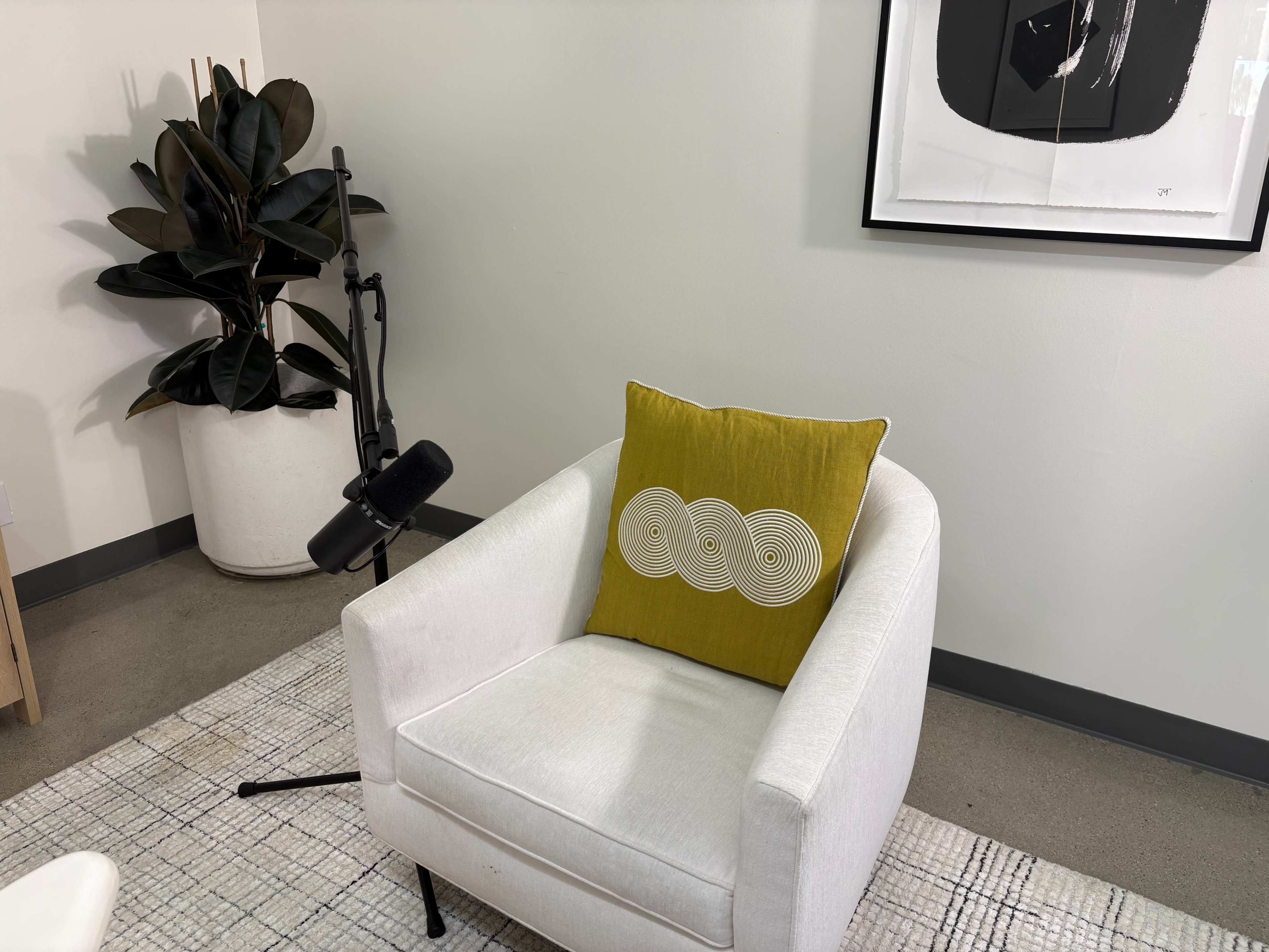 The image shows a white armchair with a yellow decorative pillow, positioned next to a potted plant and a microphone on a stand in a modern indoor space.