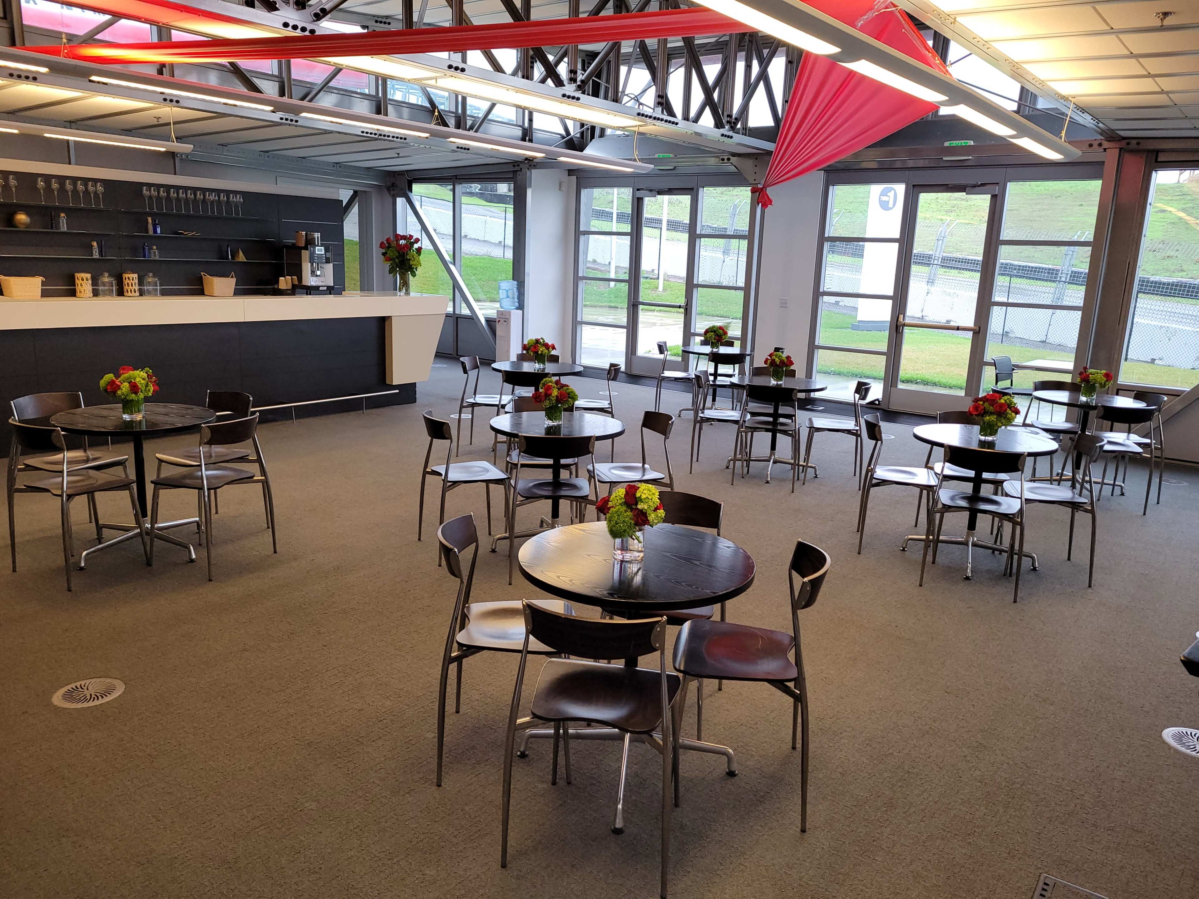 A modern indoor space with several round tables and floral centerpieces, surrounded by large windows that provide natural light.