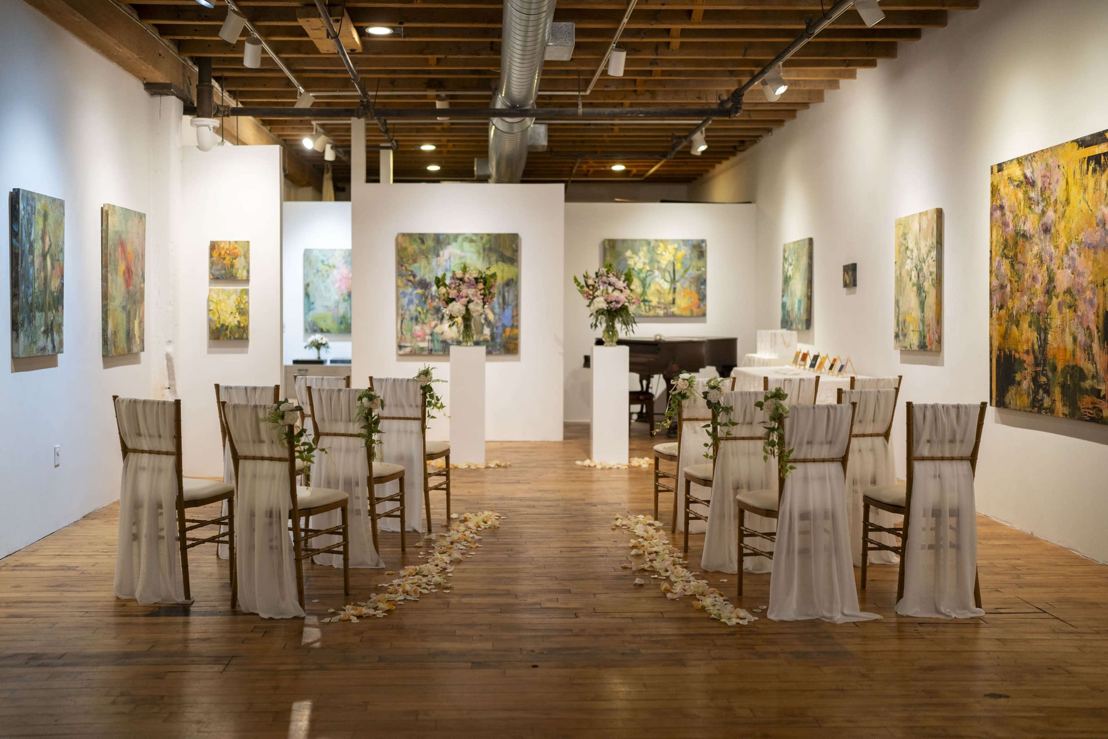 A wedding ceremony set up in an art gallery features rows of chairs adorned with white fabric and floral decorations, leading to a floral arrangement on a pedestal.