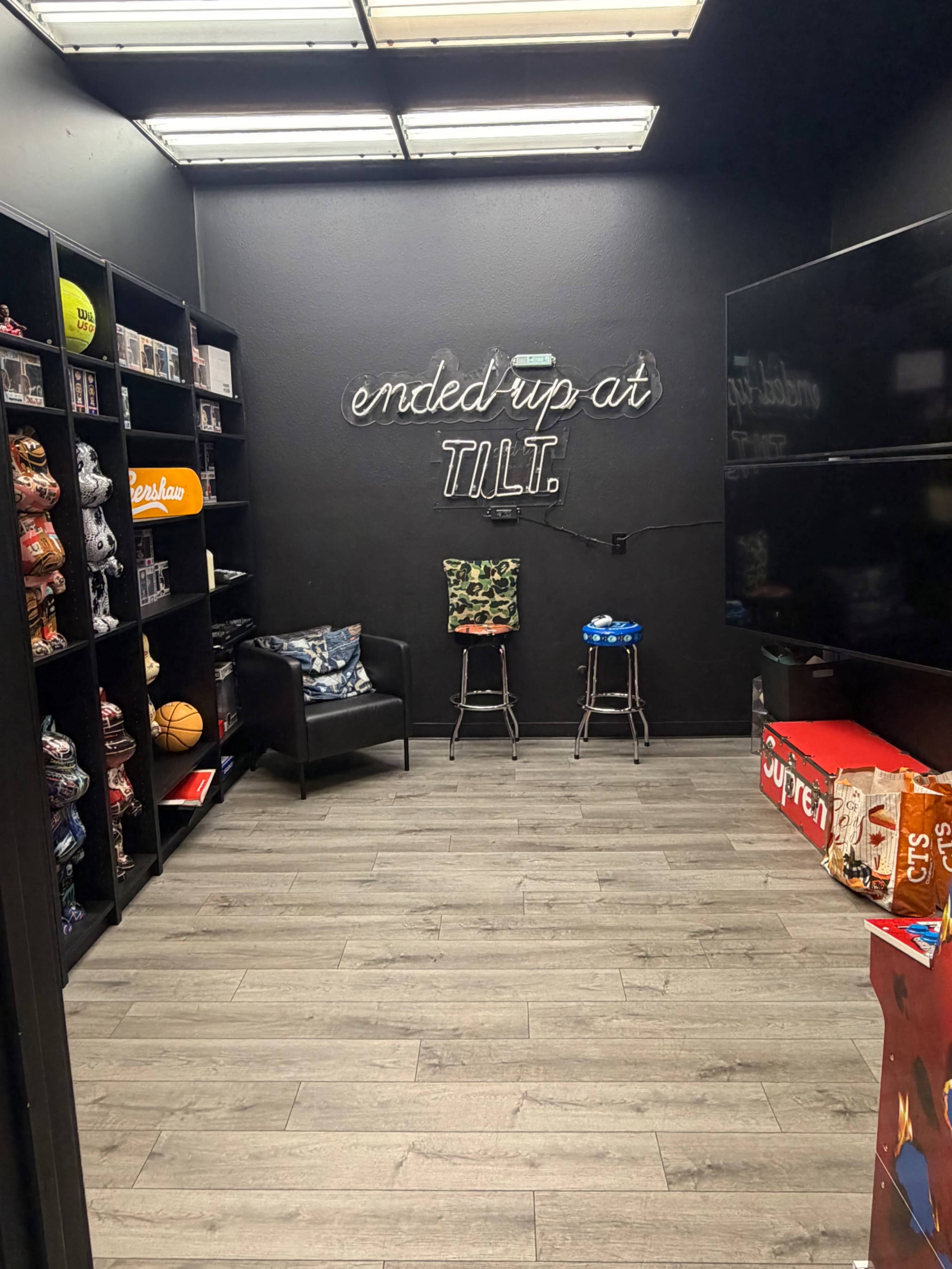 The image shows a small, darkly lit room with shelves displaying various merchandise, a pair of stools, and a TV on the wall opposite a couch.