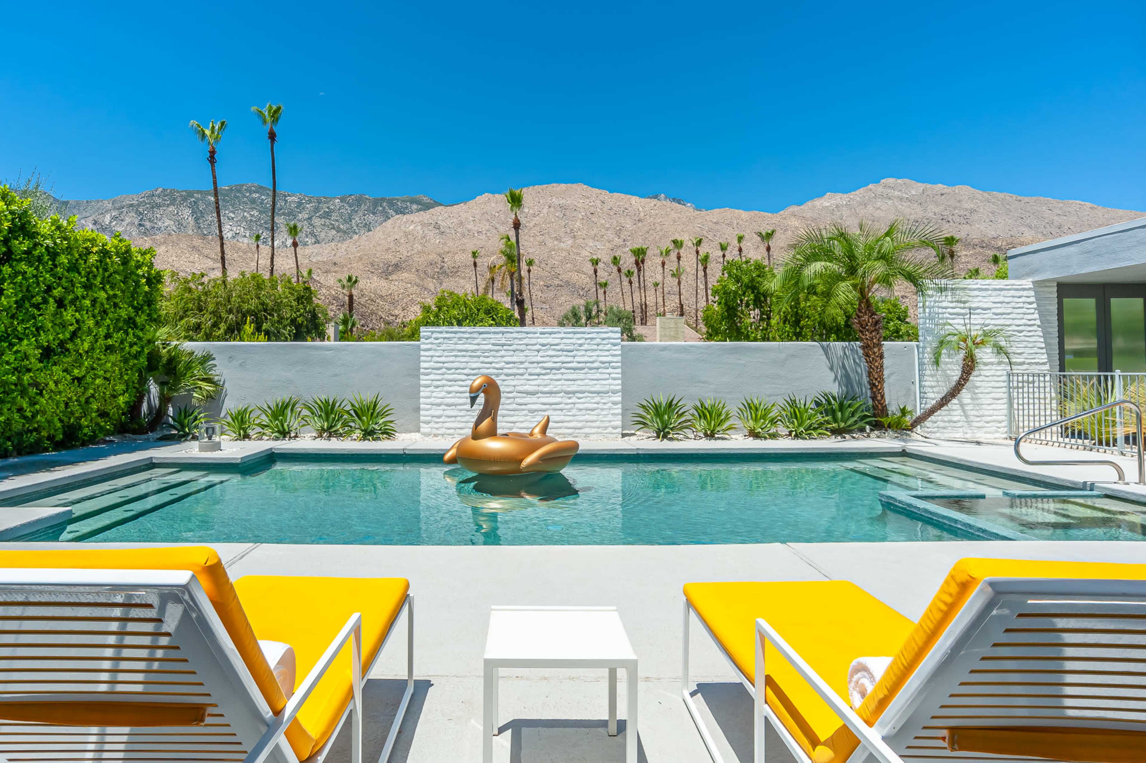 A swimming pool with a large inflatable swan is surrounded by lounge chairs and set against a backdrop of mountains and palm trees.