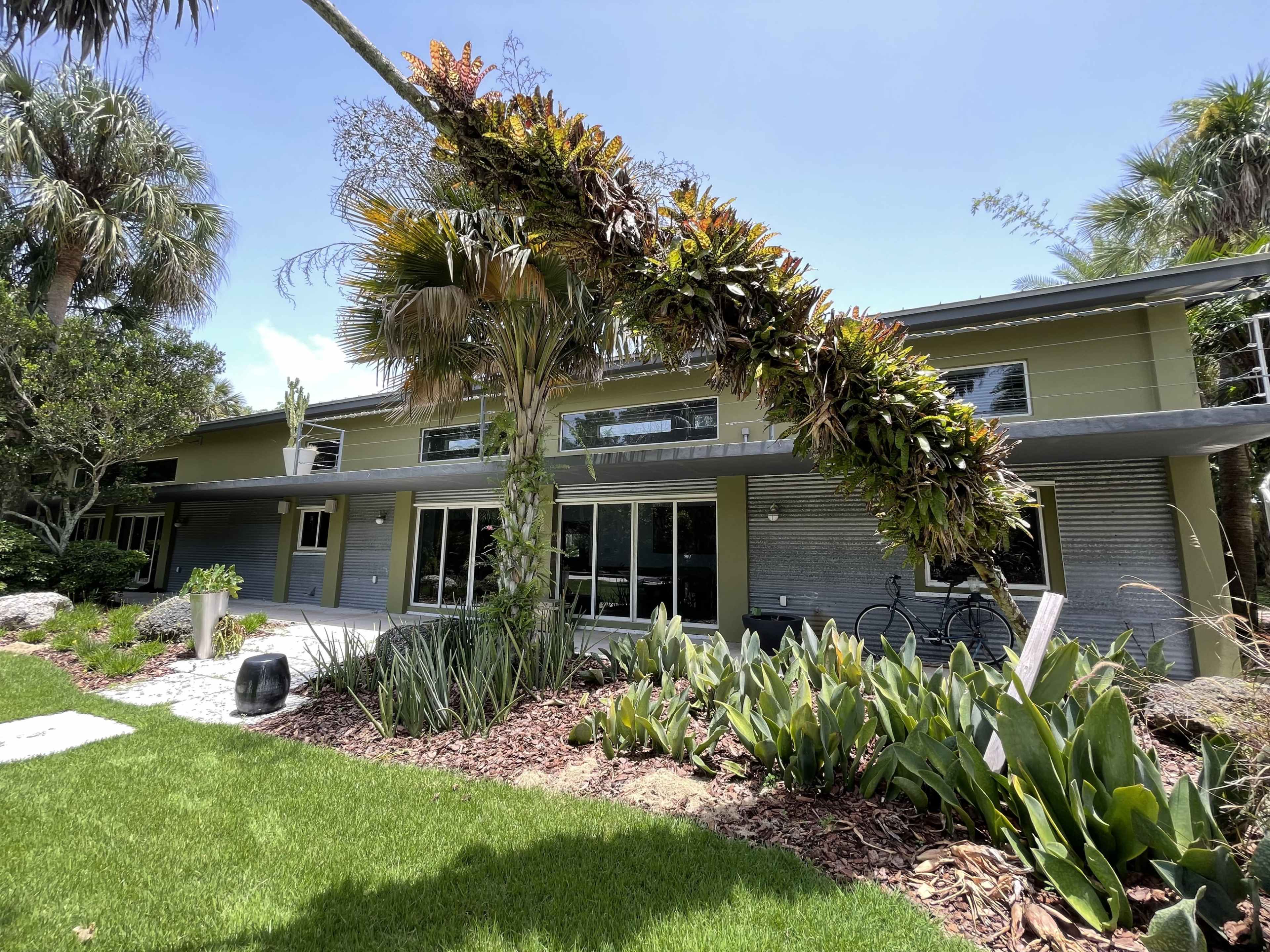 A modern house with large windows surrounded by tropical plants and landscaping.
