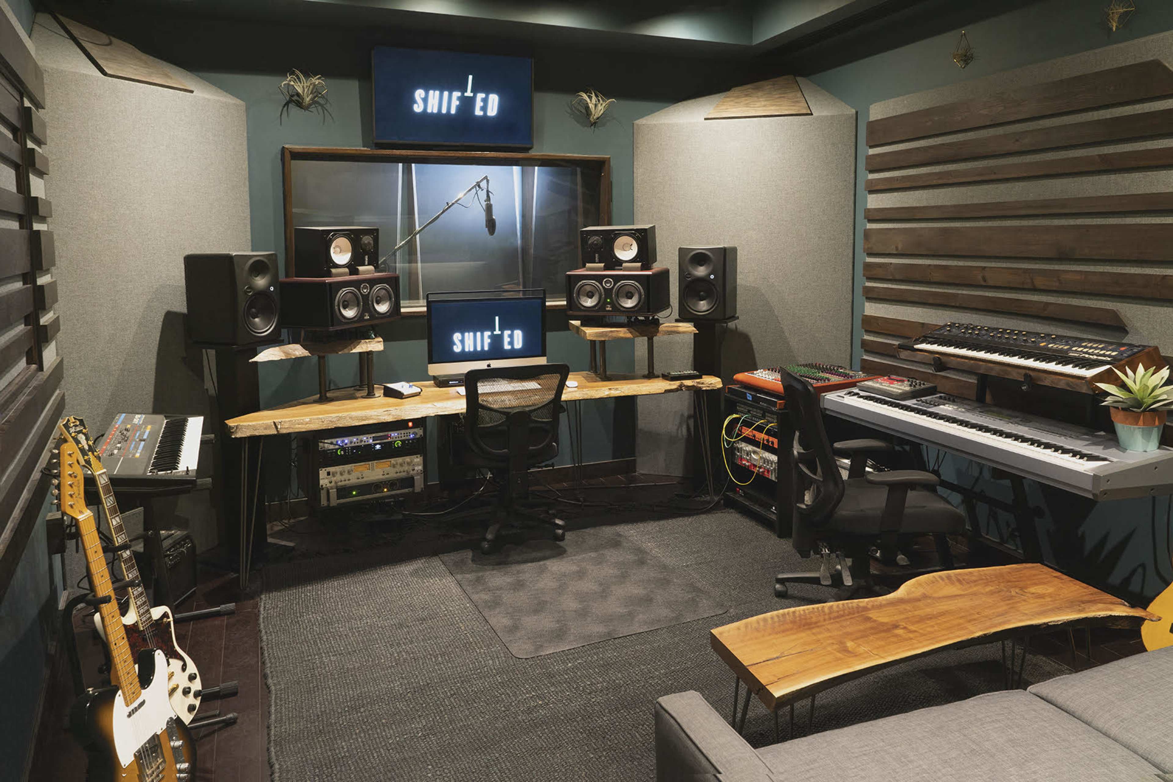 The image shows a small music production studio with a microphone setup, speakers, a keyboard, guitars, and a seating area.