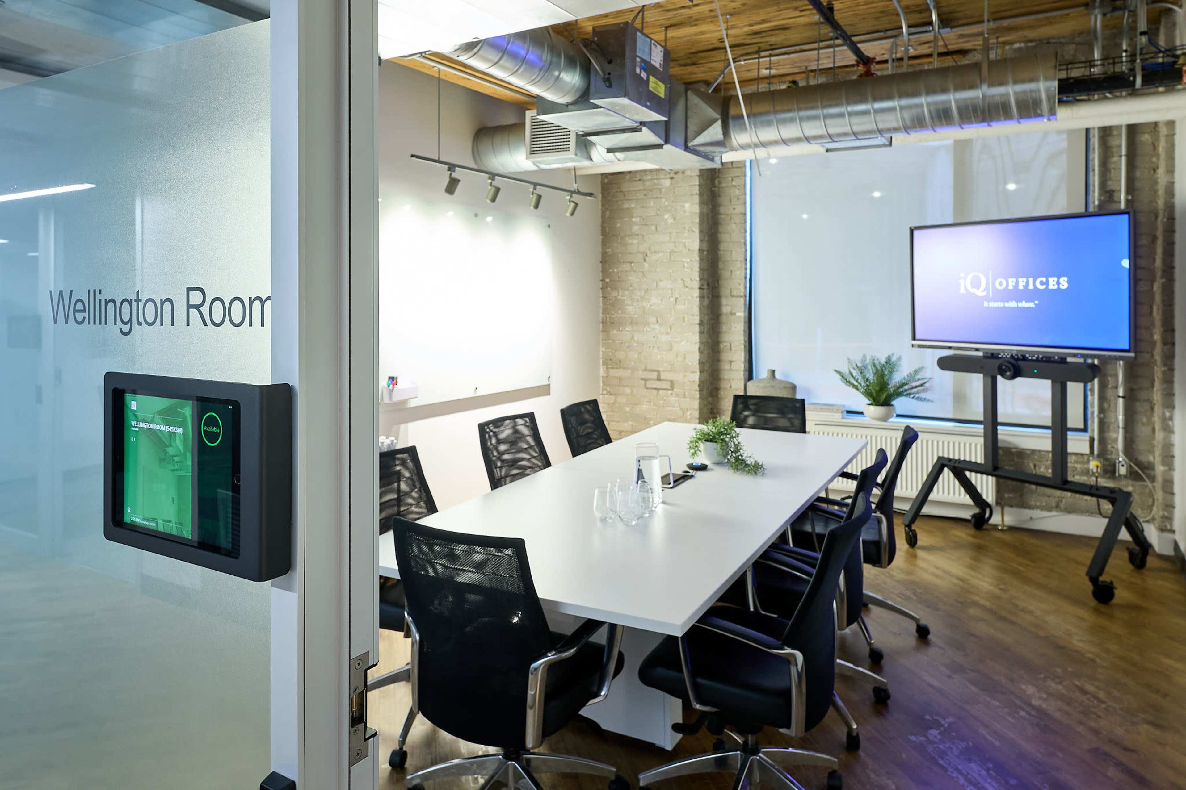 The image shows a modern conference room with a long white table, black chairs, and a large screen displaying the "iQ Offices" logo.
