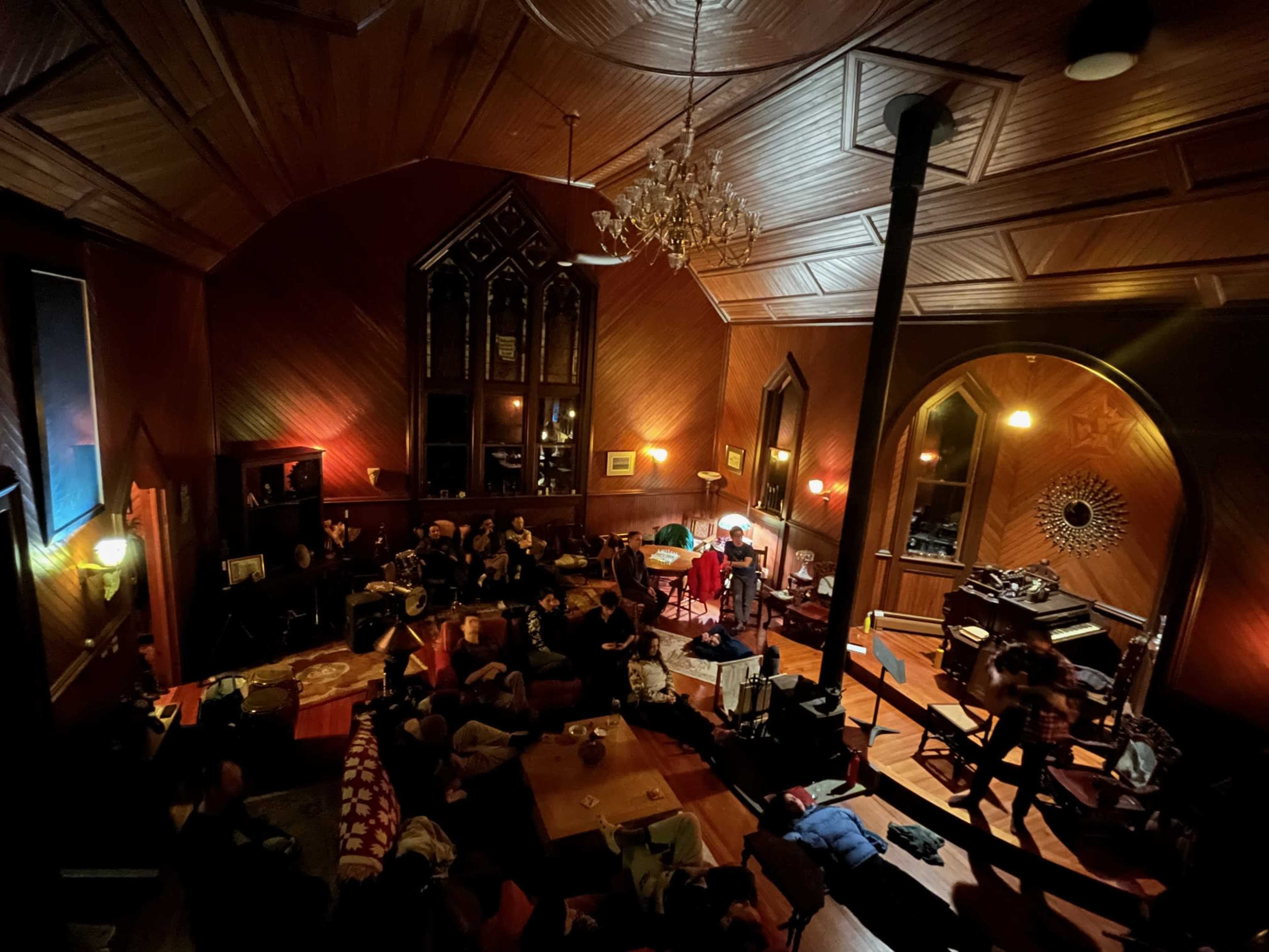 A spacious wooden room with high ceilings, where a group of people is gathered, surrounded by furniture and musical instruments.