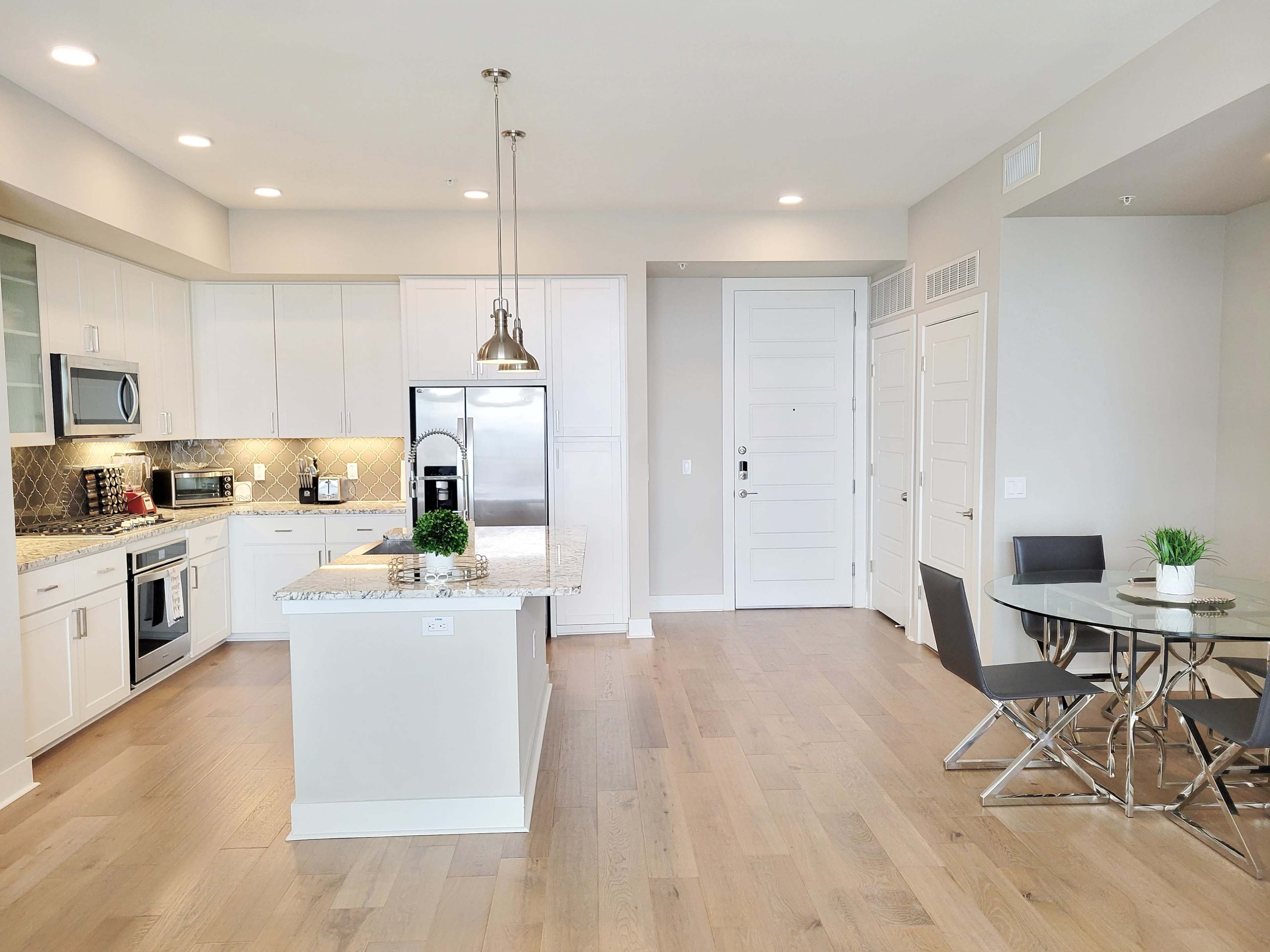 A modern kitchen and dining area featuring white cabinetry, stainless steel appliances, a granite countertop island, and a small glass dining table with metal chairs.