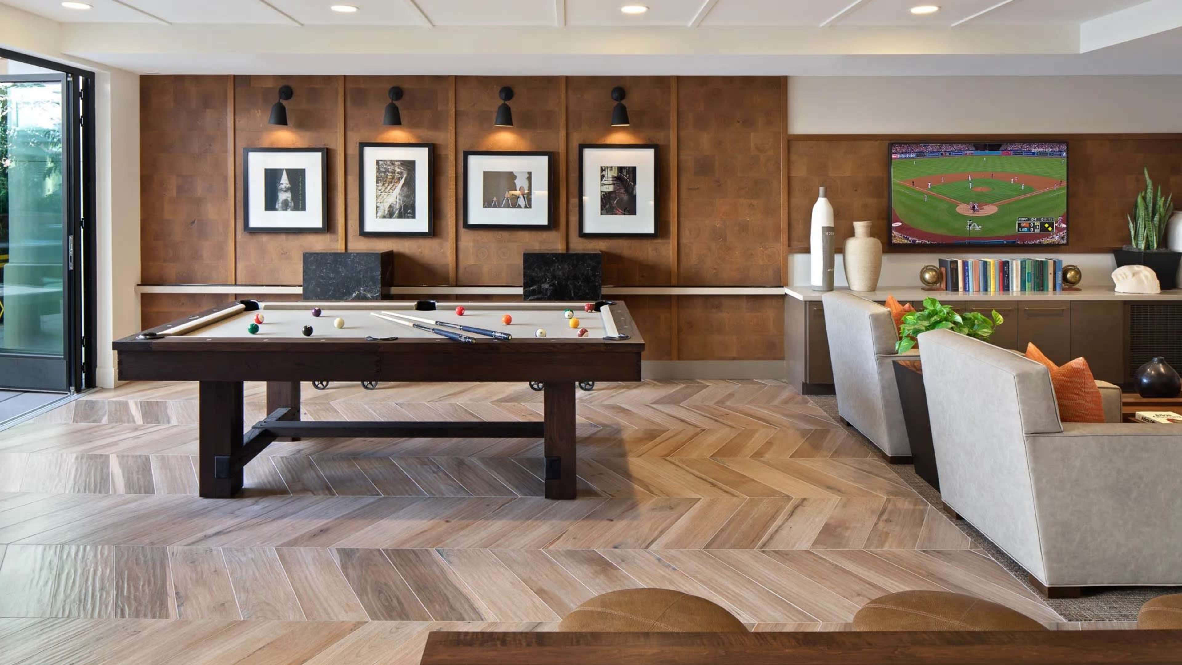 A modern game room featuring a pool table in the center, framed artworks on the wall, a television broadcasting a sports event, and a comfortable seating area.