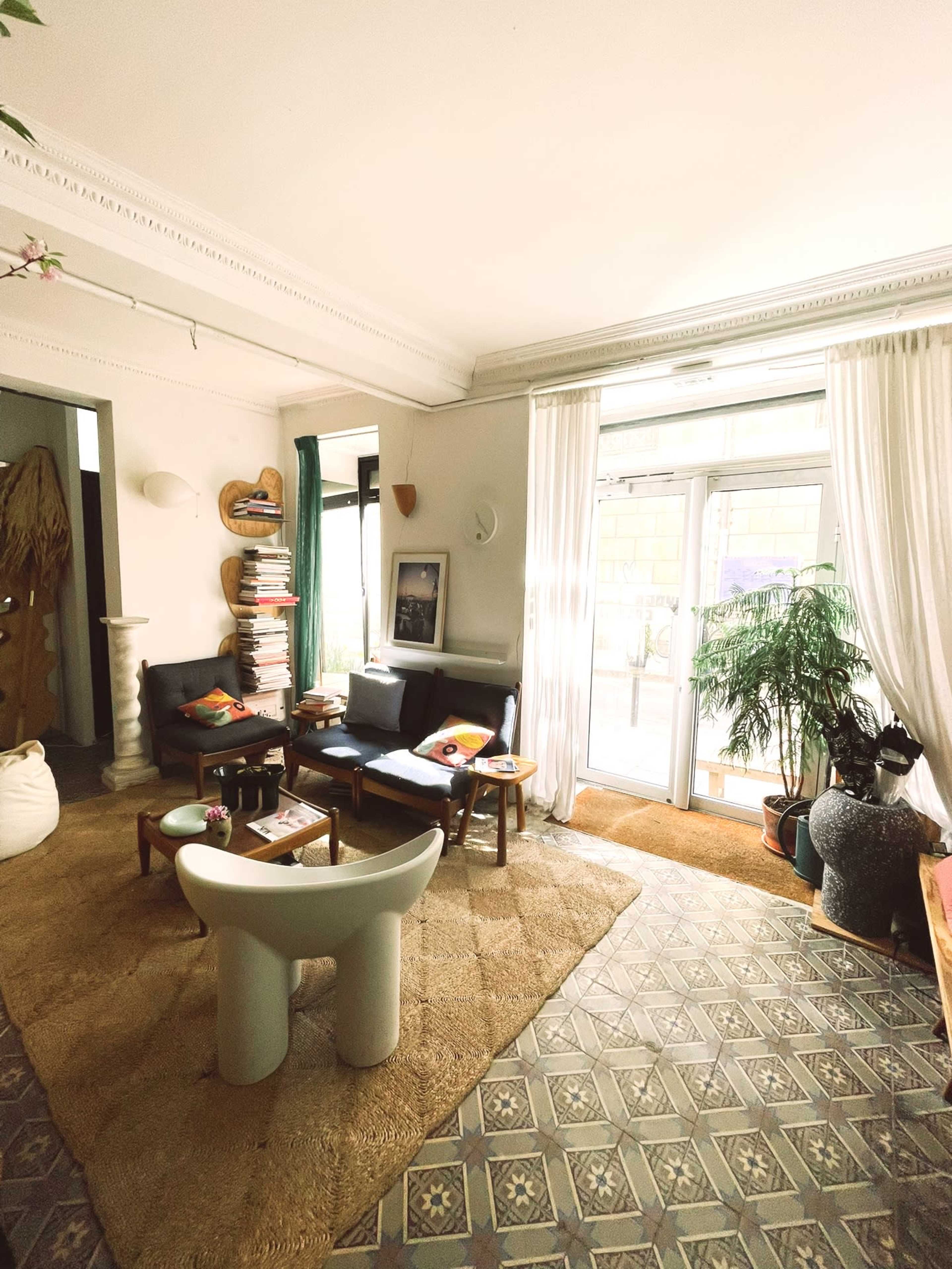 A cozy living room with a seating area, a large window allowing natural light, and plants adorning the space.