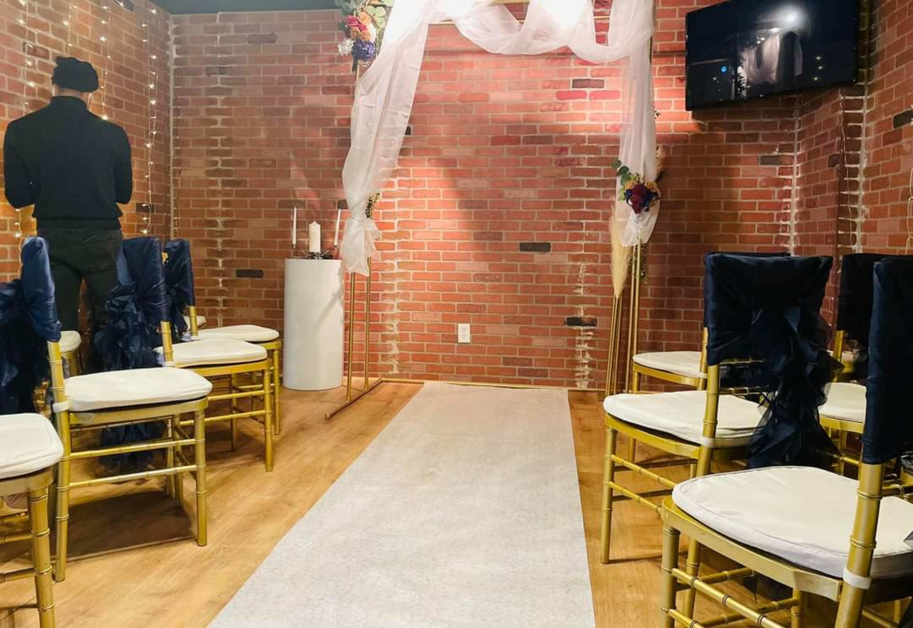 A small indoor ceremony space features rows of gold chairs with navy blue sashes facing a white aisle, adorned with fabric drapery and a small altar against a brick wall.