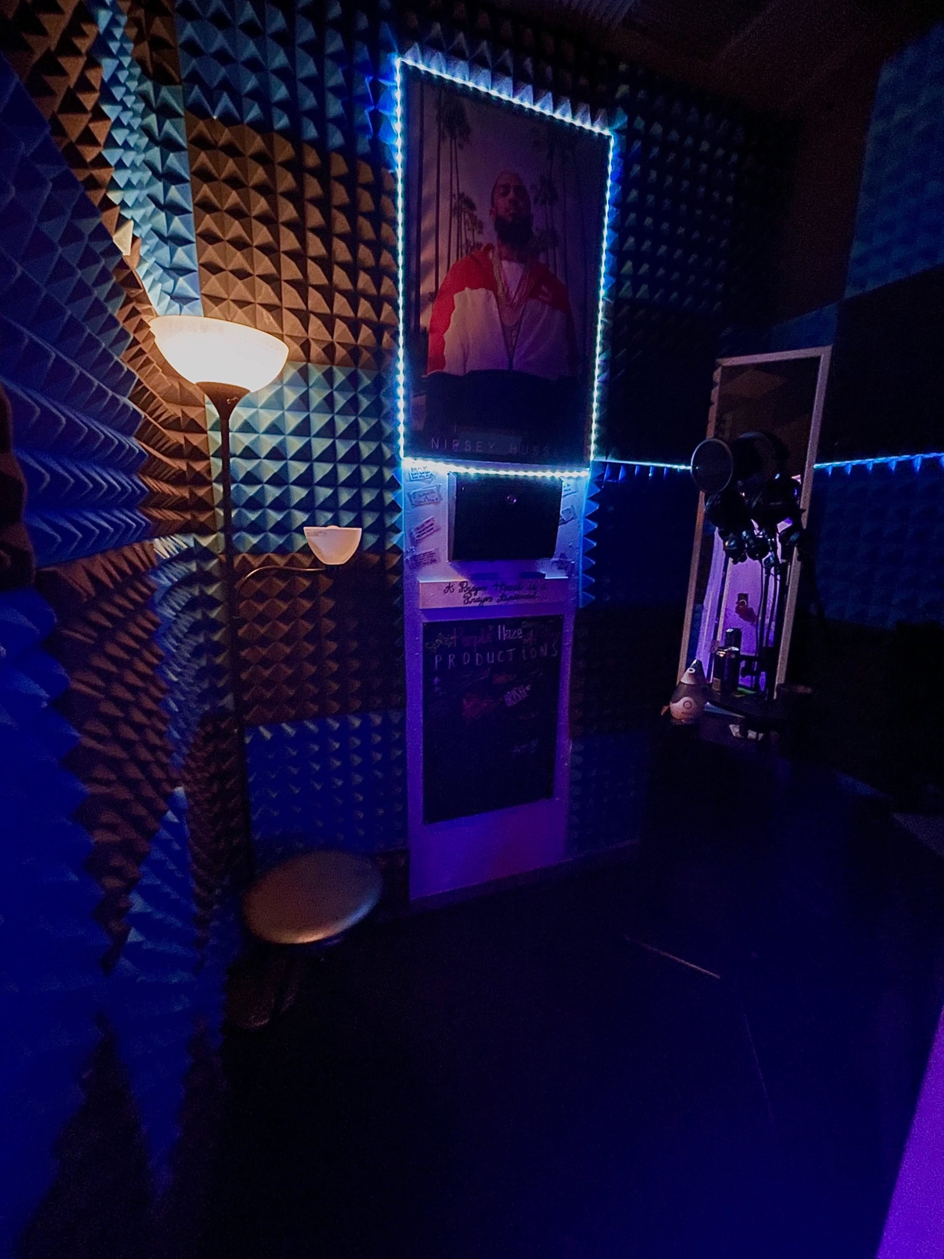 The image shows a small, soundproofed room with acoustic foam walls, a lamp, and a framed poster on the wall.