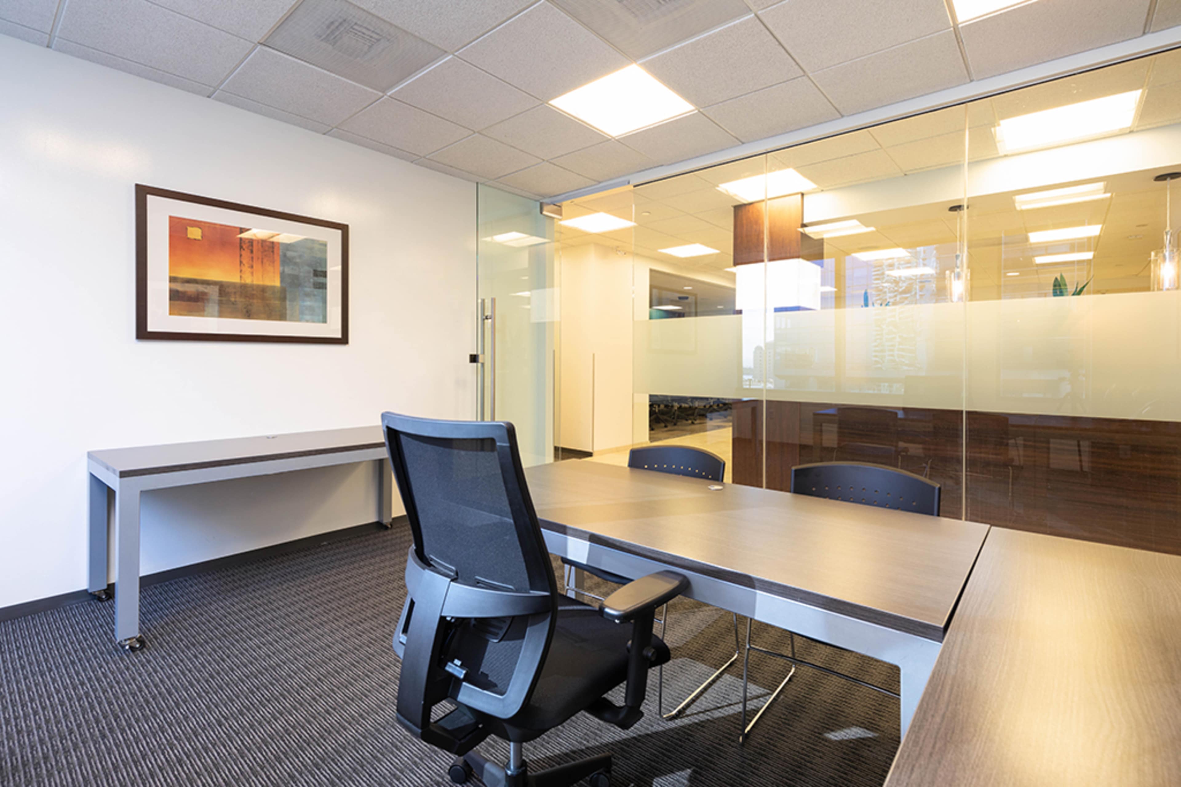 An empty office space features a desk, an office chair, and a glass-walled conference room in the background.