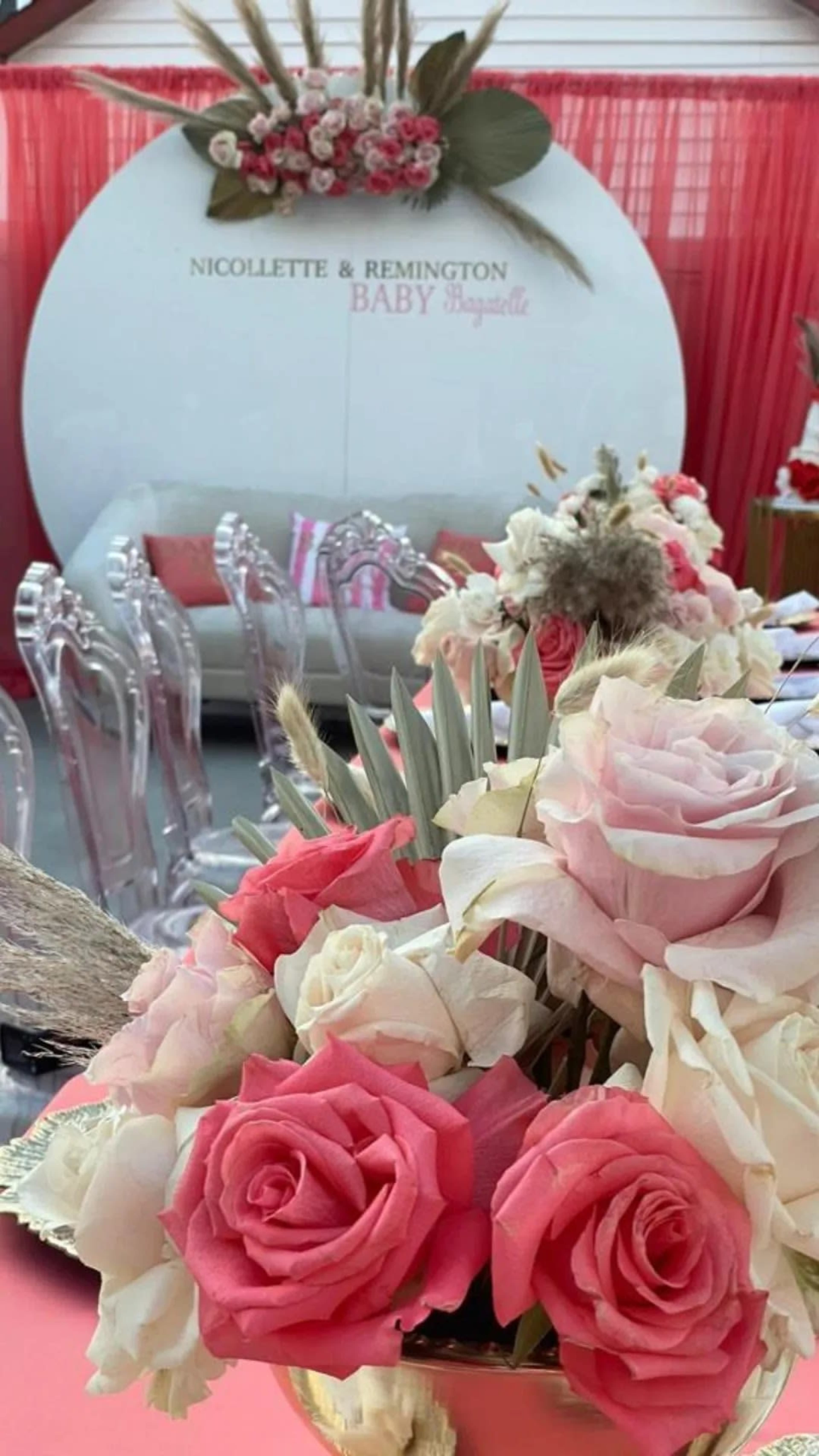 The image features a floral arrangement in the foreground with pink and cream roses, while a background displays a decorated area for a baby celebration, including a circular sign with the names "NICOLETTE & REMINGTON" and a seating area.