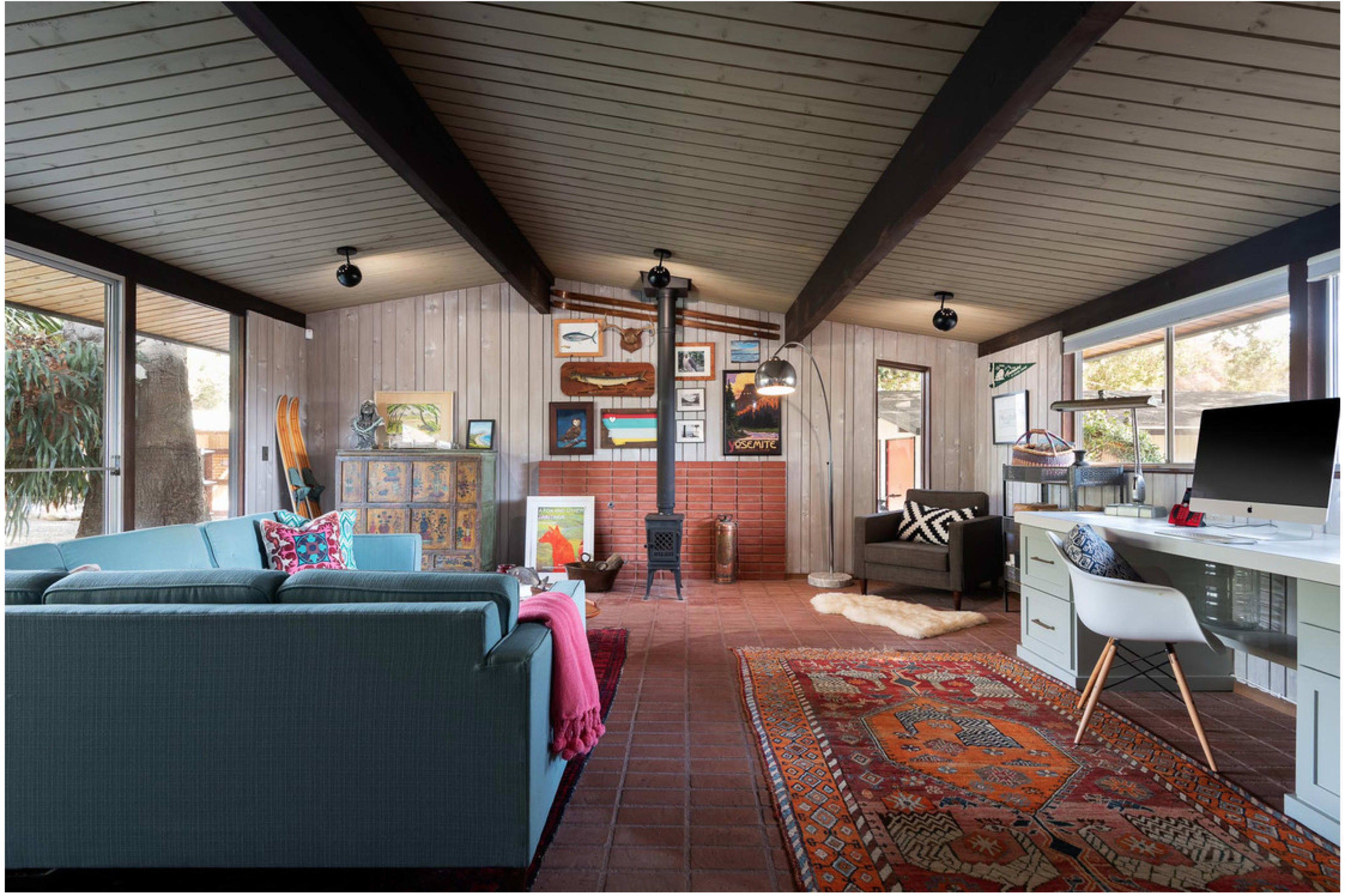 The image shows a cozy living space with a mix of modern and rustic decor, featuring a blue couch, a wooden stove, a desk, and various artworks on the walls.