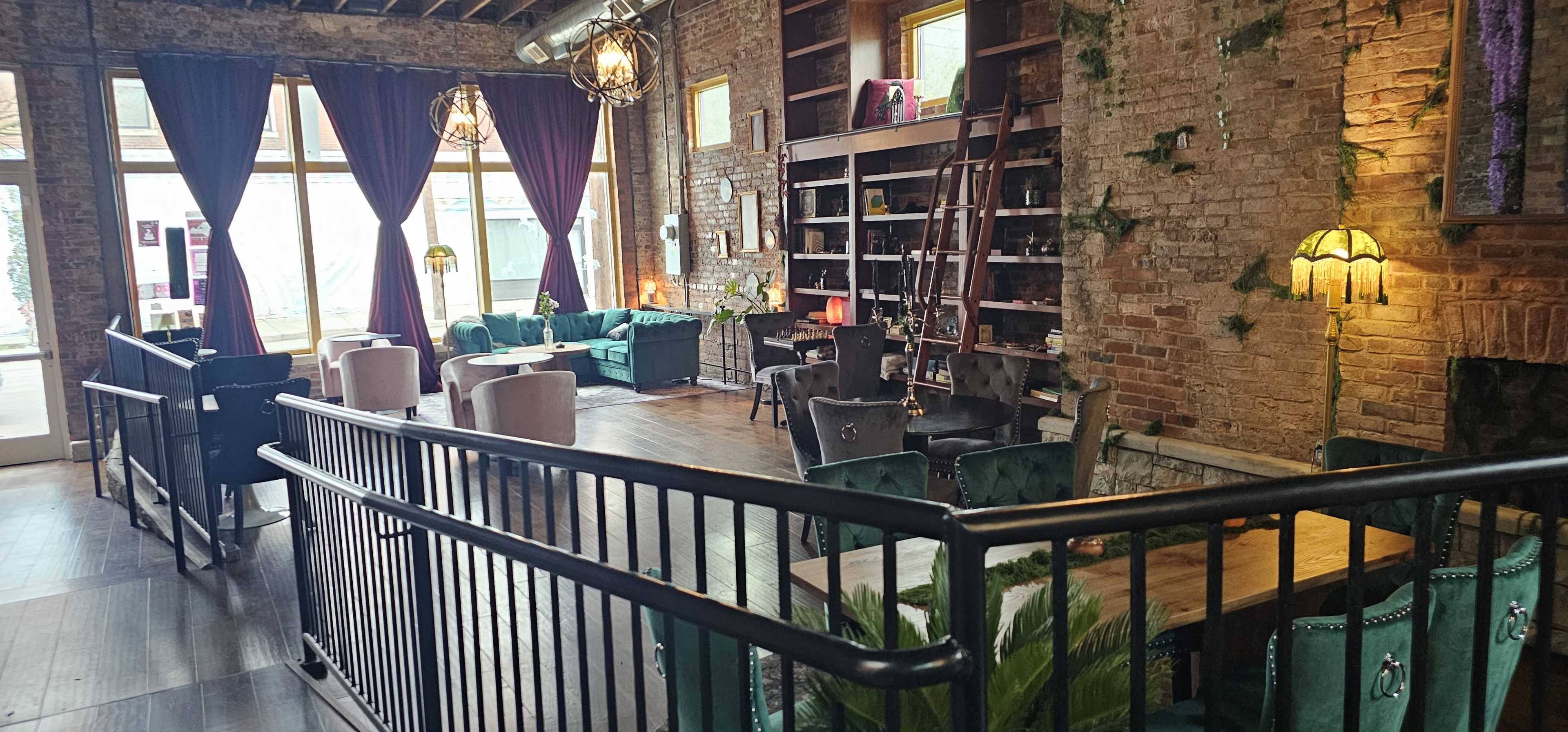 The image shows a cozy interior of a cafe or lounge with brick walls, plush green seating, and decorative lighting, separated by a black railing.
