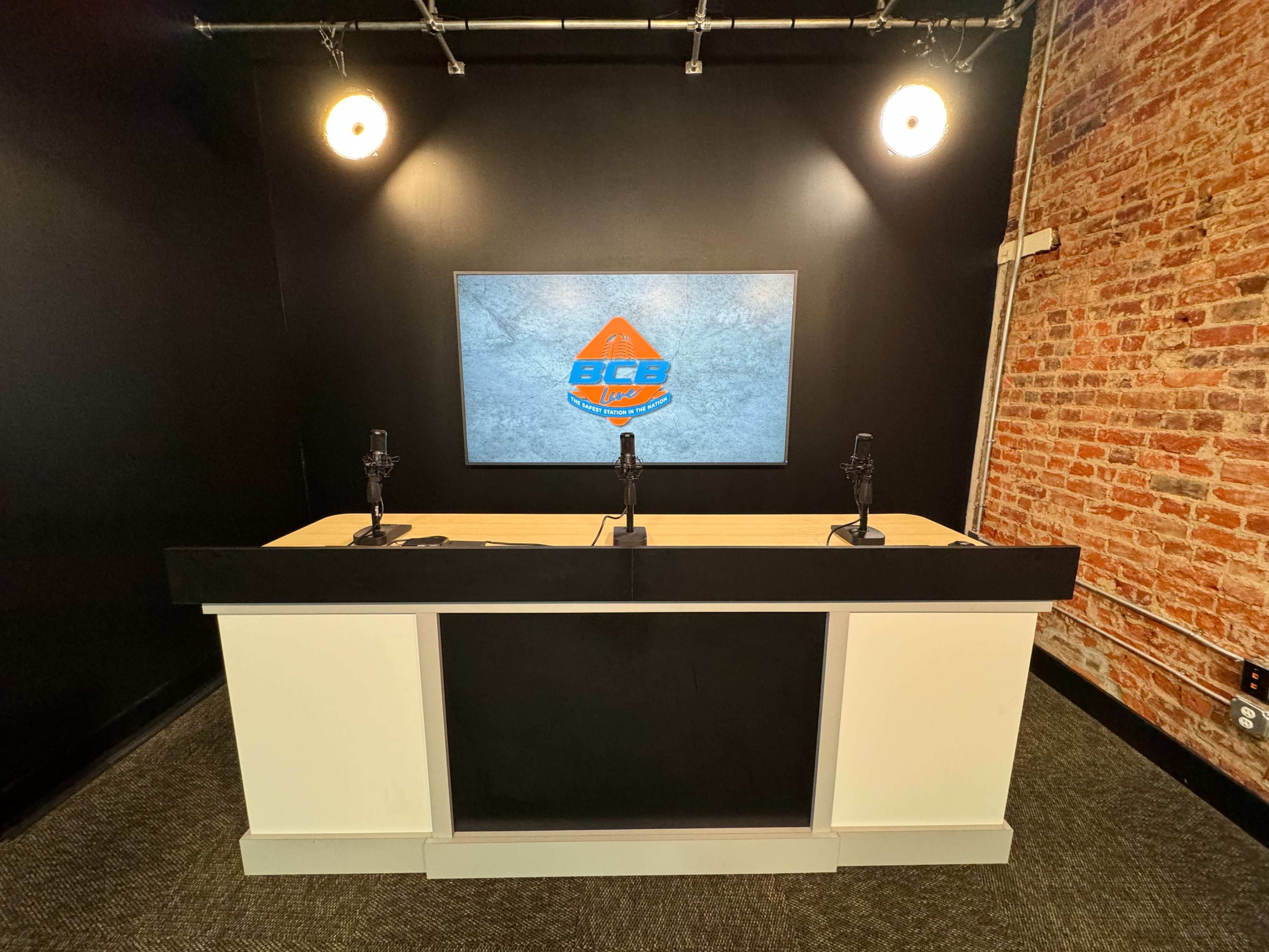 A modern broadcasting studio with a black and white counter, two microphones, and a large screen displaying a logo against a brick wall.