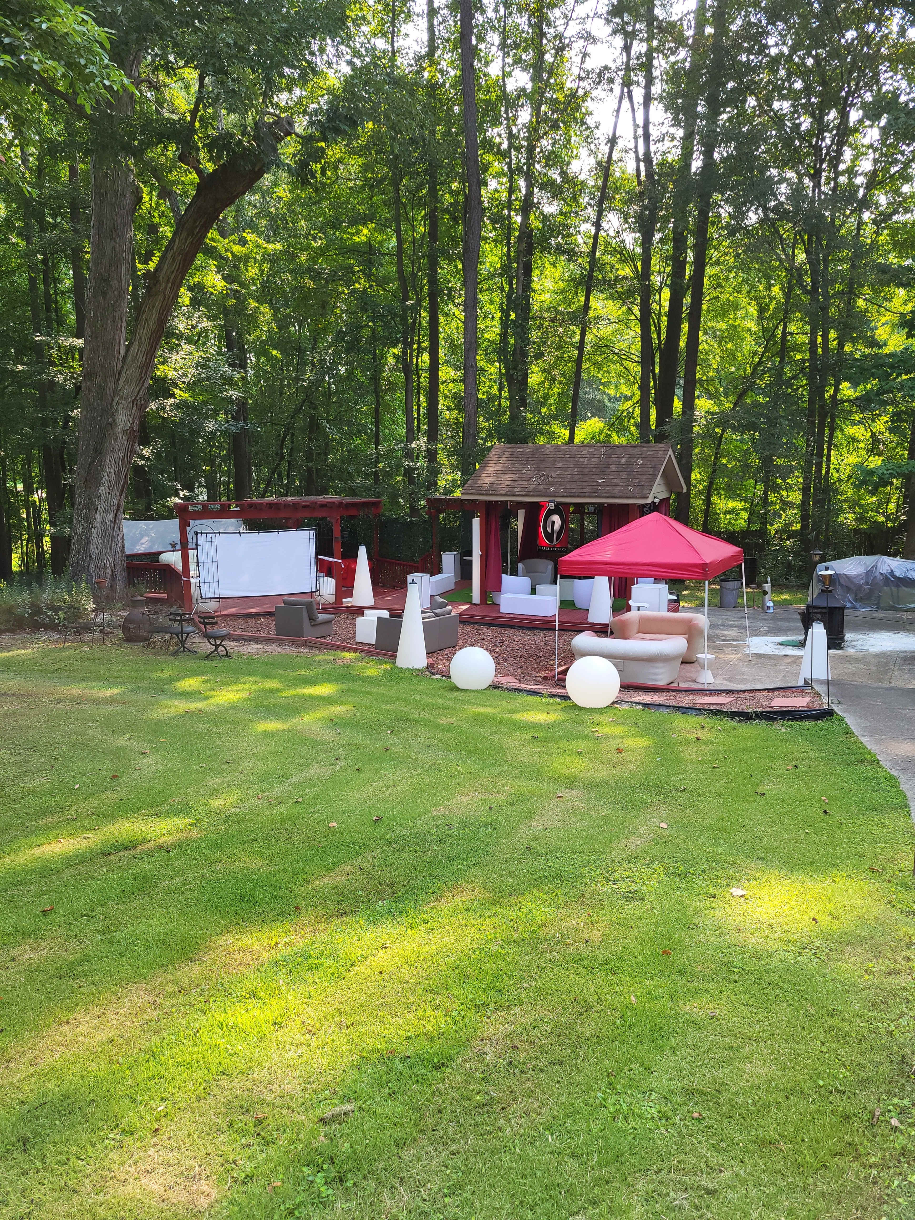 The image shows a landscaped outdoor area featuring a seating arrangement under a red umbrella, a small hut, and a large screen among trees.