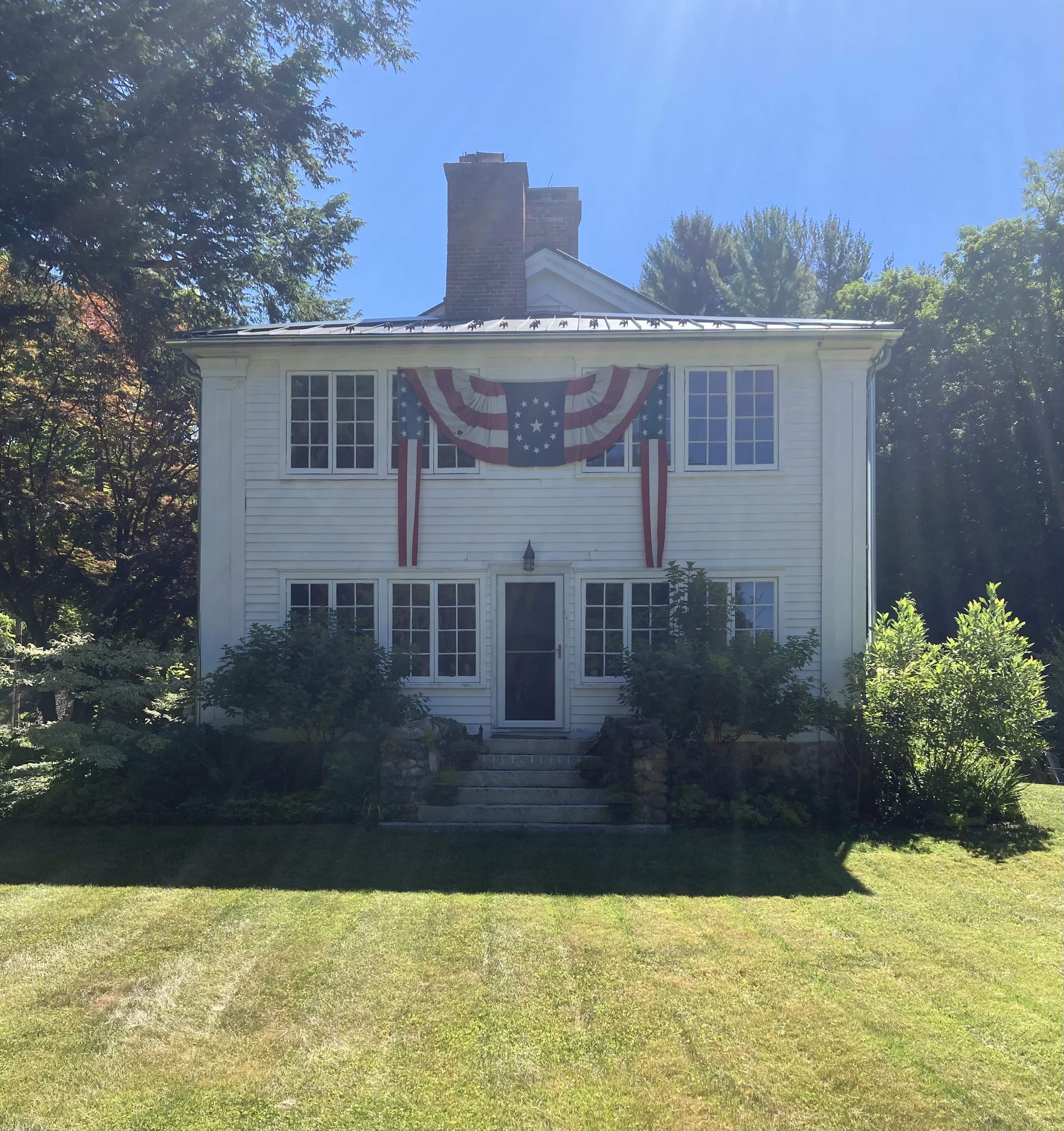 A white two-story house with a front porch is adorned with red, white, and blue decorations, set in a grassy yard under clear blue skies.