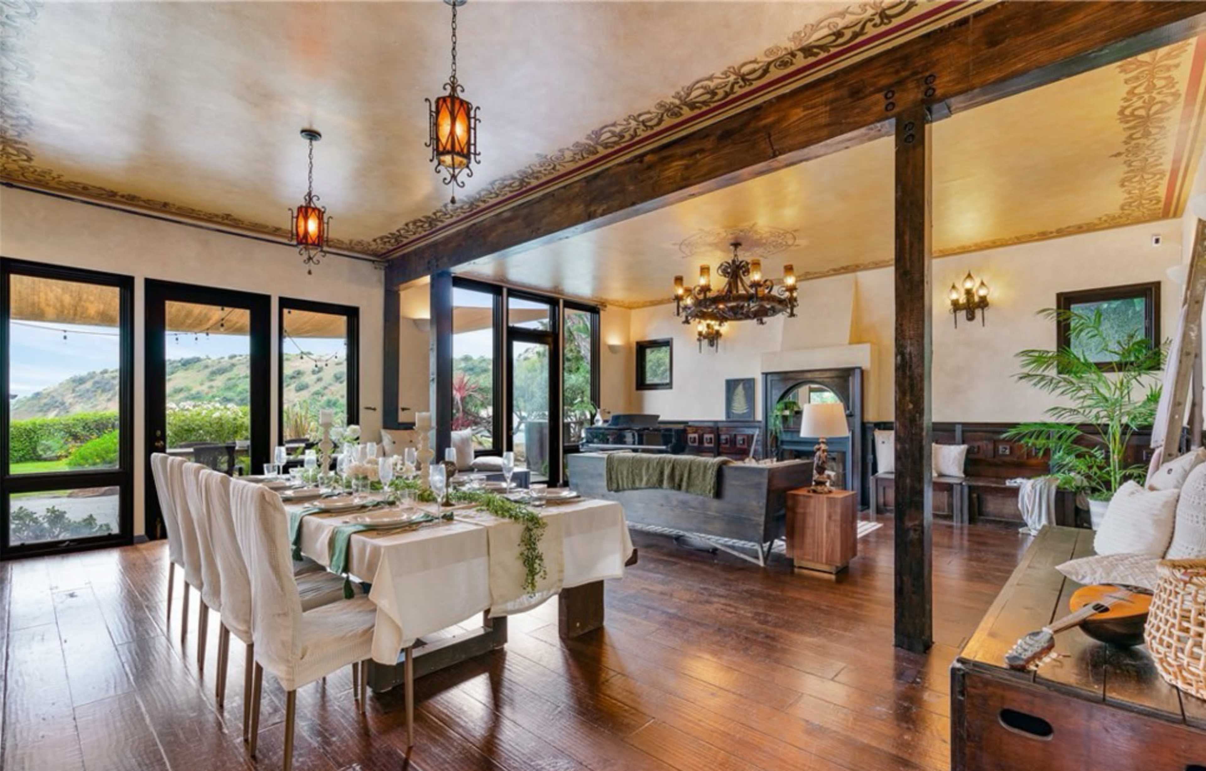The image shows a spacious dining area with a long table set for a meal, surrounded by large windows that offer a view of the outdoors.