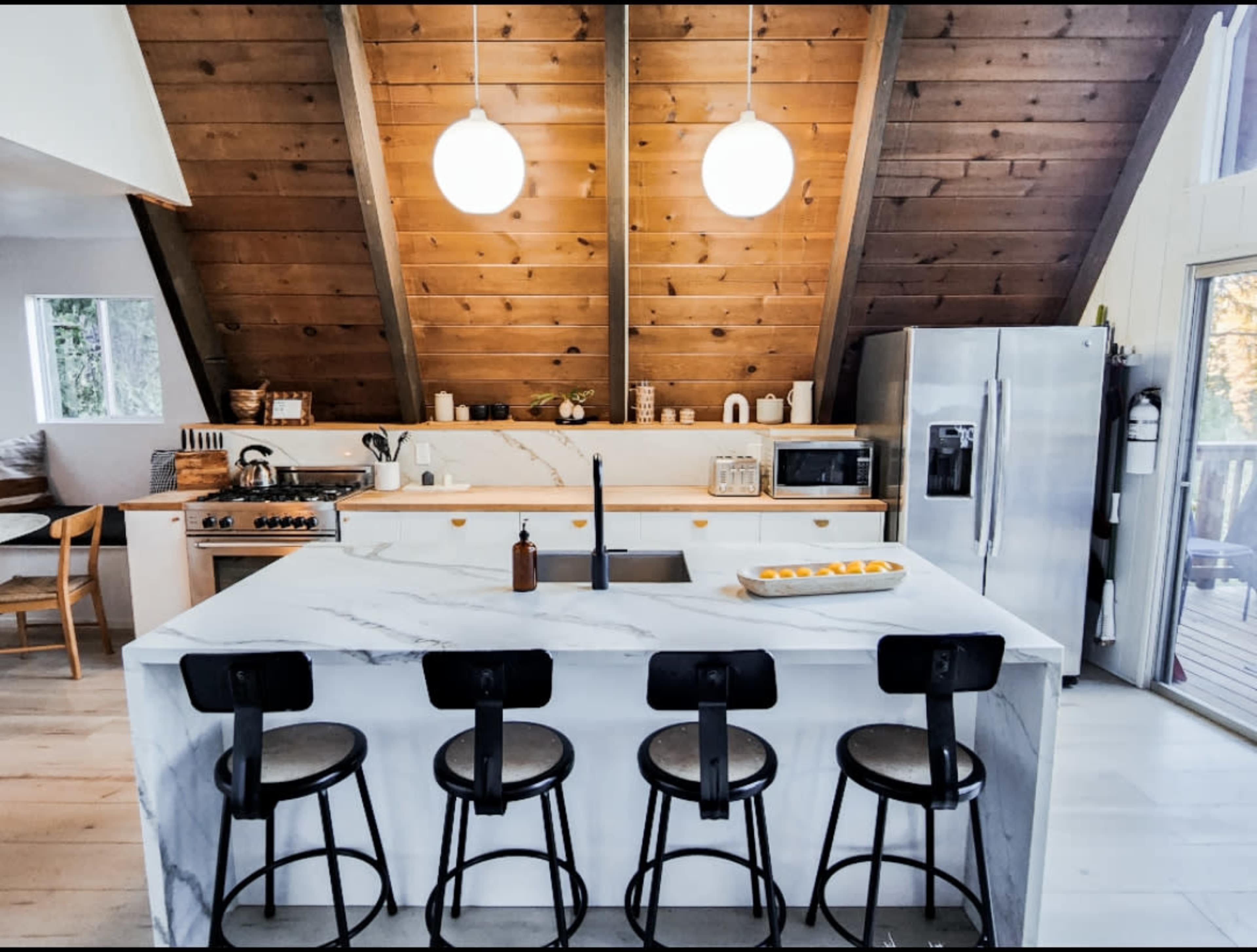 The image shows a modern kitchen with a marble island, four black stools, and a wooden sloped ceiling.
