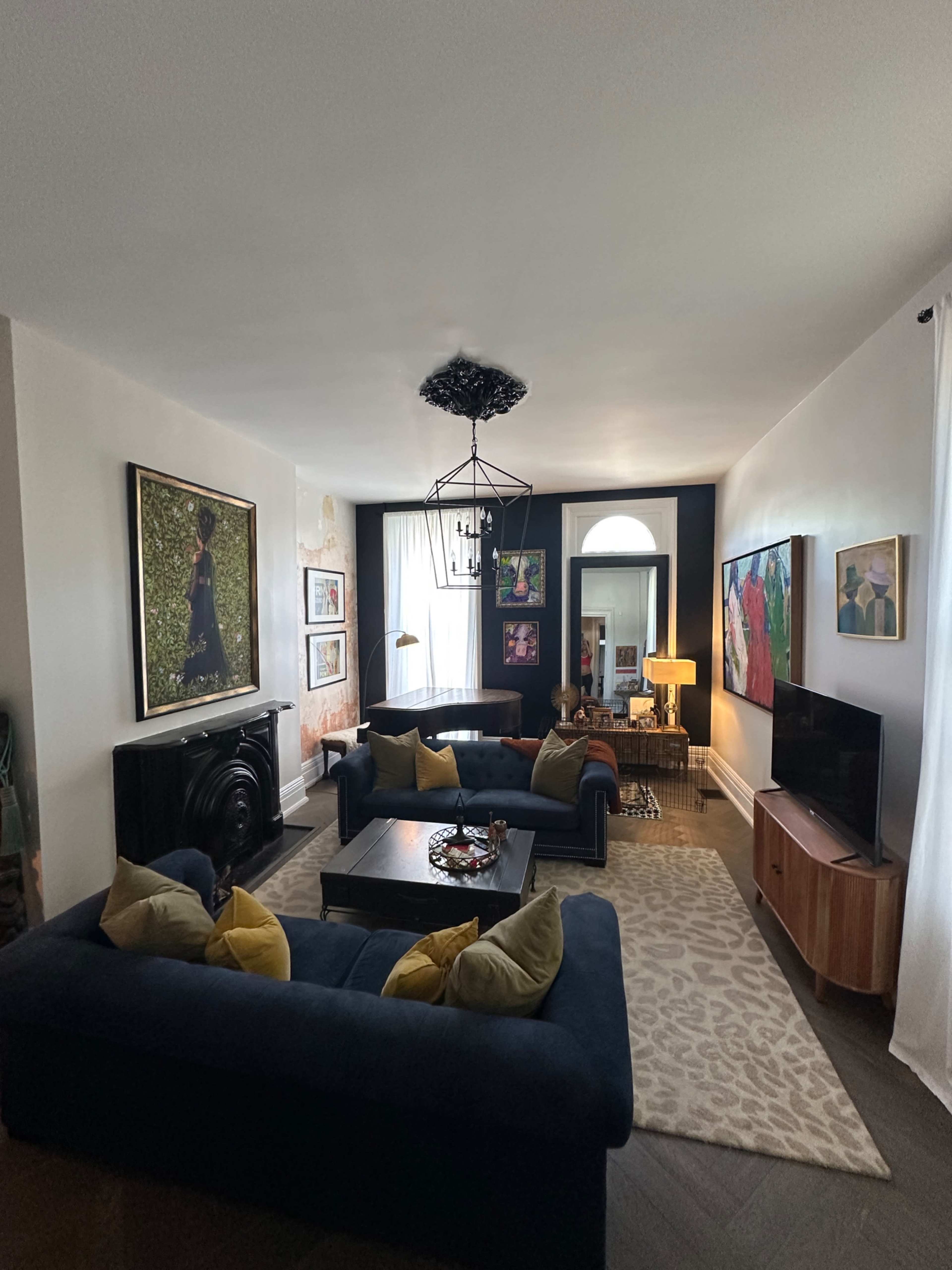 The image shows a modern living room featuring two dark blue sofas, a coffee table, and a flat-screen TV, with various artworks on the walls and large windows providing natural light.