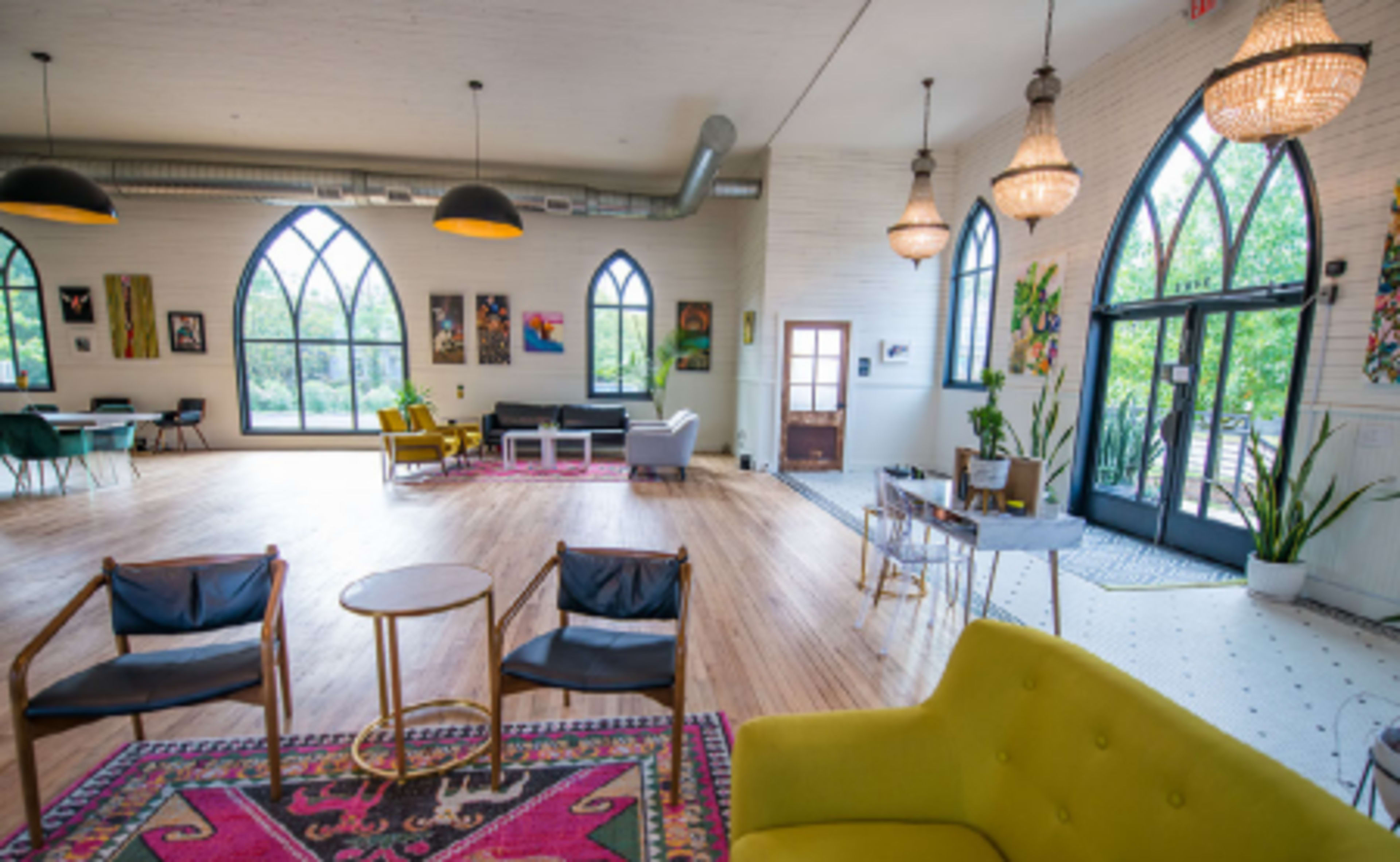 A spacious, well-lit interior with large arched windows, wooden flooring, and various seating arrangements, including chairs, tables, and artwork on the walls.