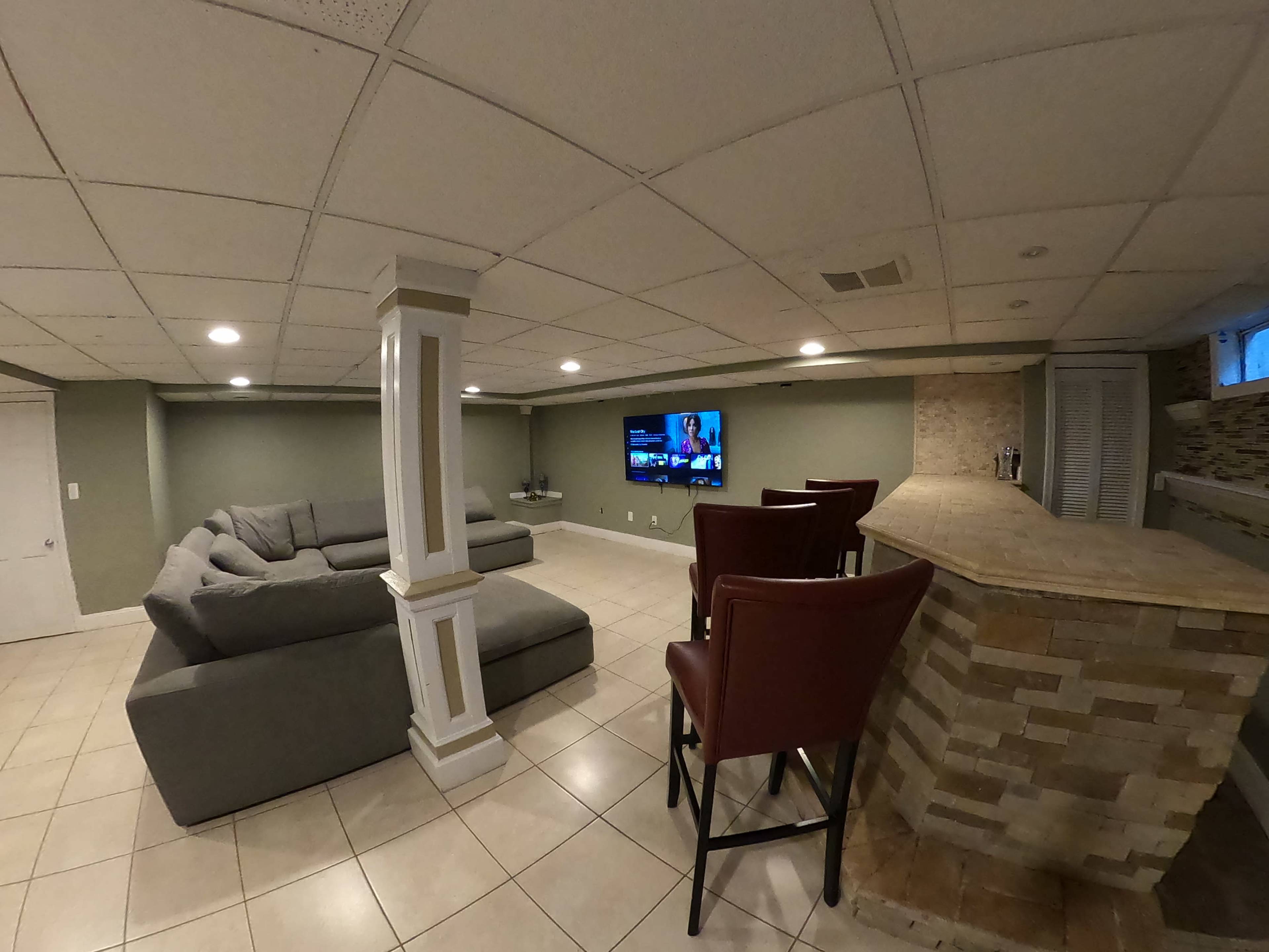 A spacious basement features a gray sectional sofa, a stone bar with red chairs, and a mounted television on the wall.