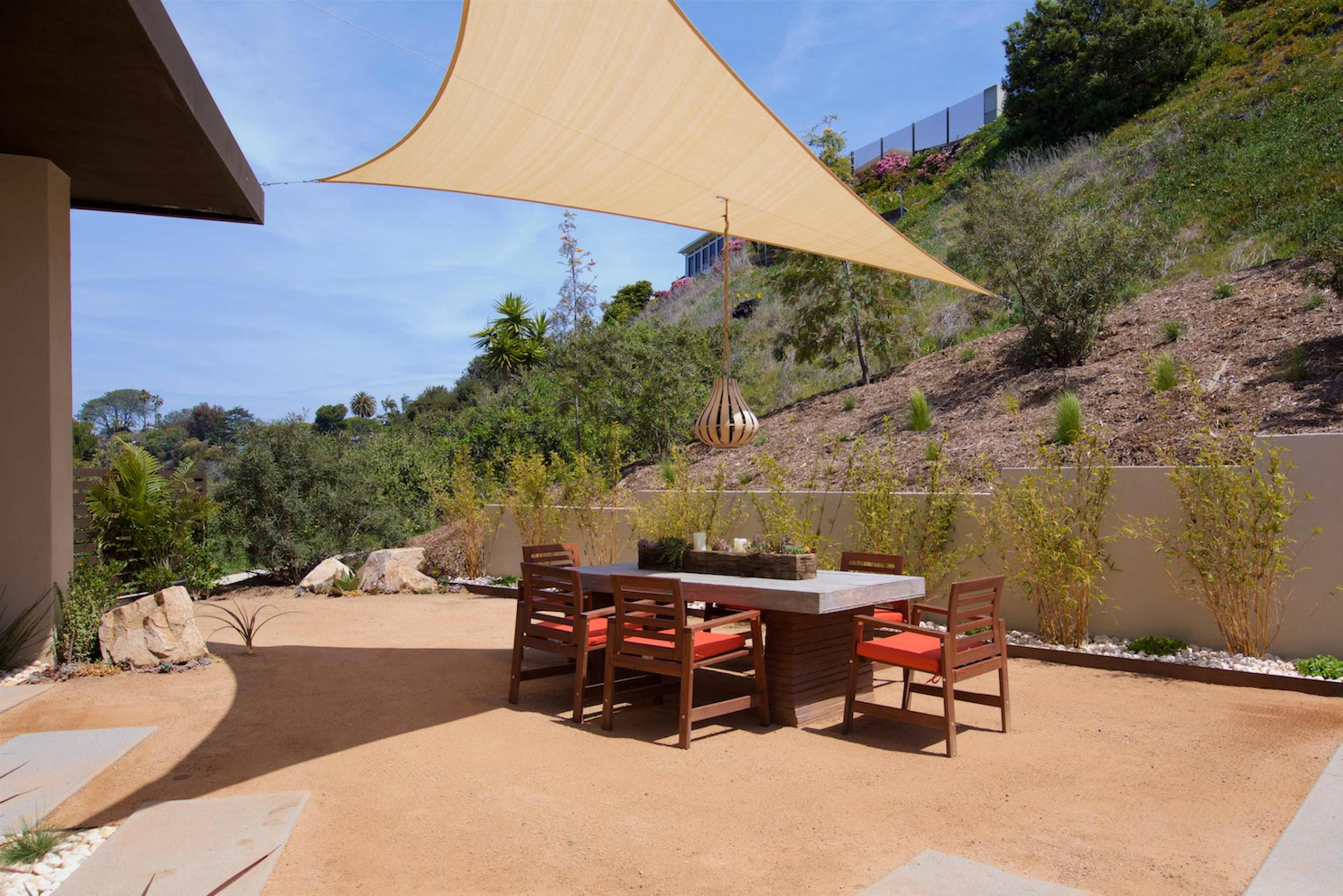 A shaded outdoor dining area features a stone table surrounded by wooden chairs on a sandy patio.