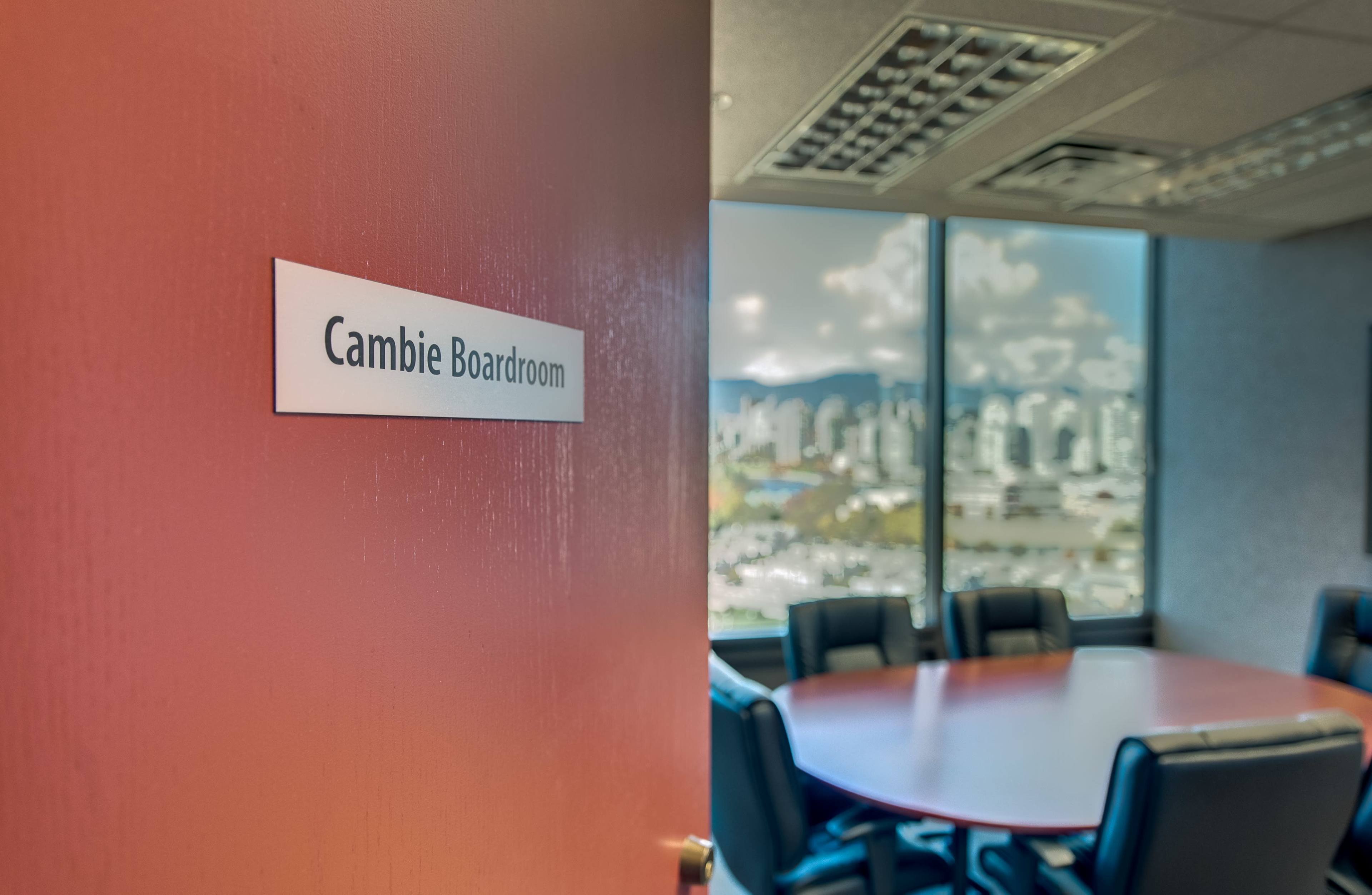 A red door with a sign reading "Cambie Boardroom" opens to a meeting room featuring a circular table and large windows showing a city skyline with mountains in the background.