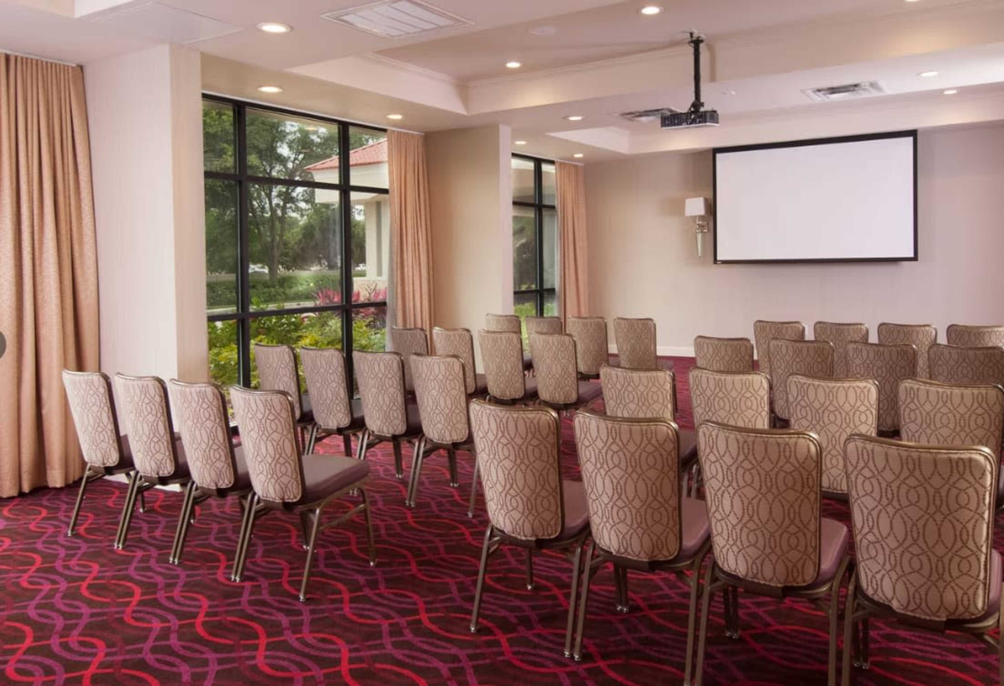 The image shows a conference room set up with rows of patterned chairs facing a blank projector screen.