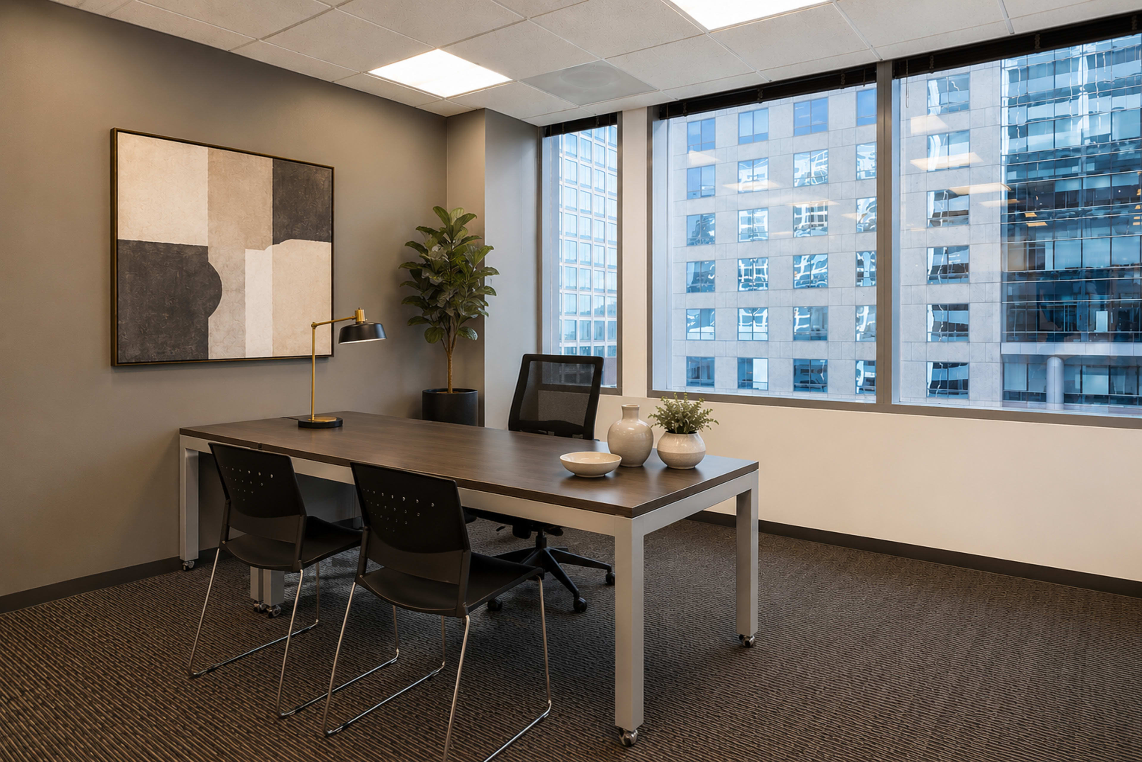 The image shows a modern office space with a large desk, two black chairs, a table lamp, and a potted plant, all positioned near large windows overlooking a cityscape.