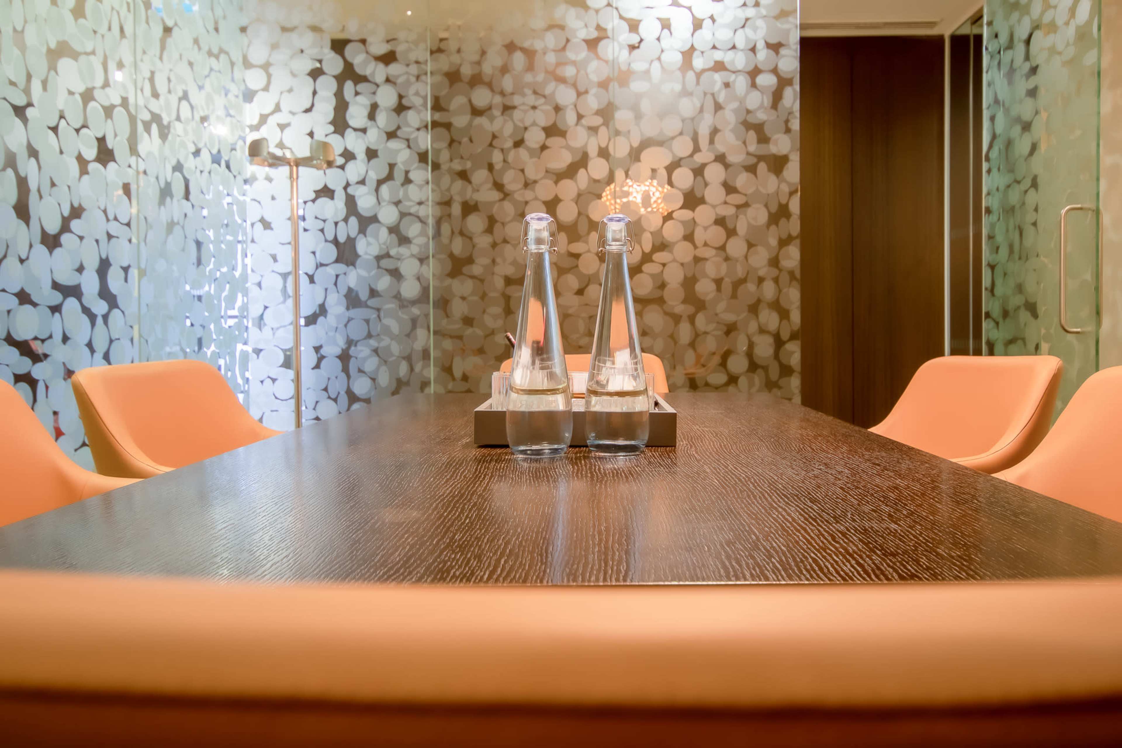 The image shows a modern conference room with a wooden table, orange chairs, and two bottles of water on a tray.
