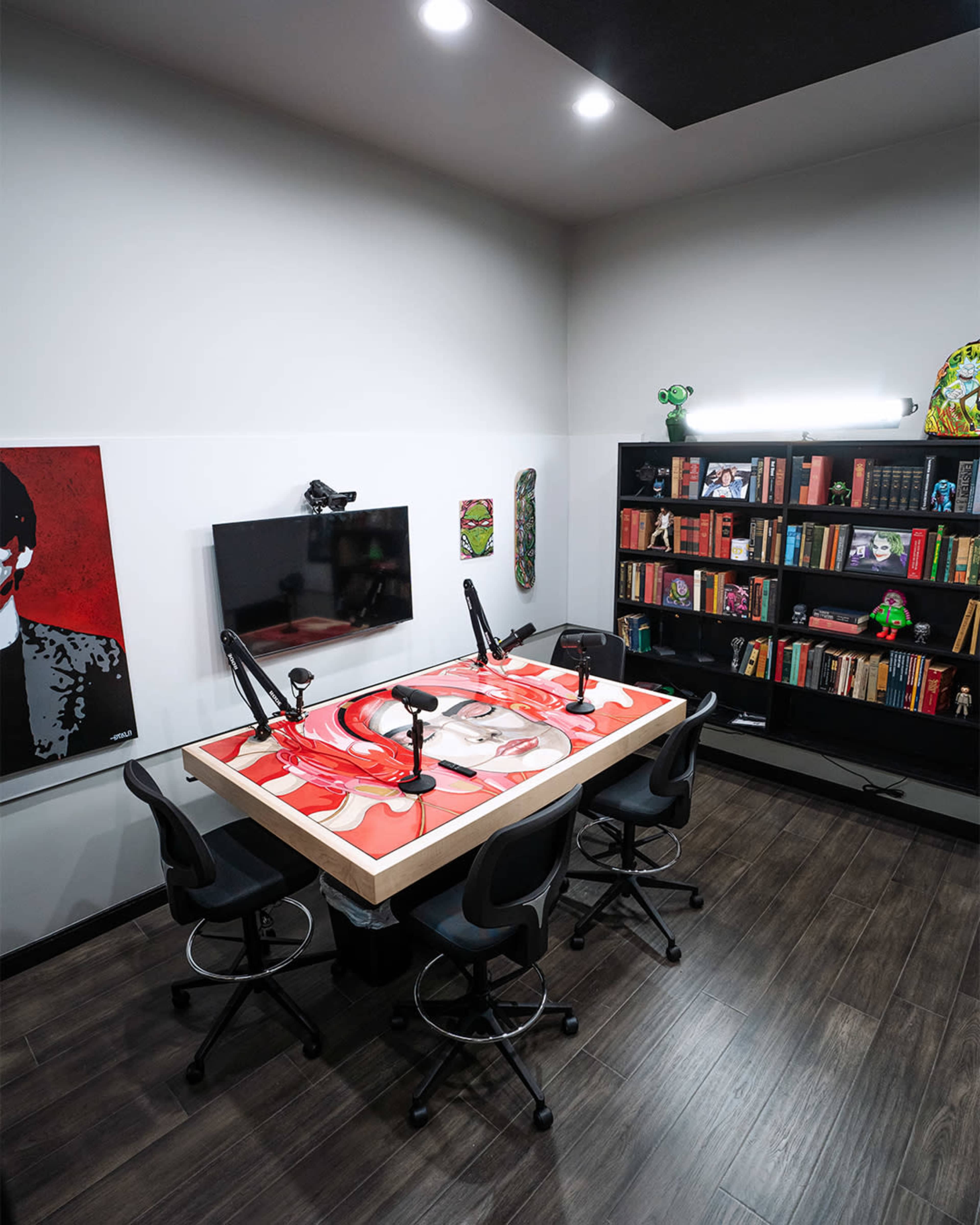 A modern podcasting studio featuring a table with four microphones, a television mounted on the wall, and a bookshelf filled with books and decorative items.