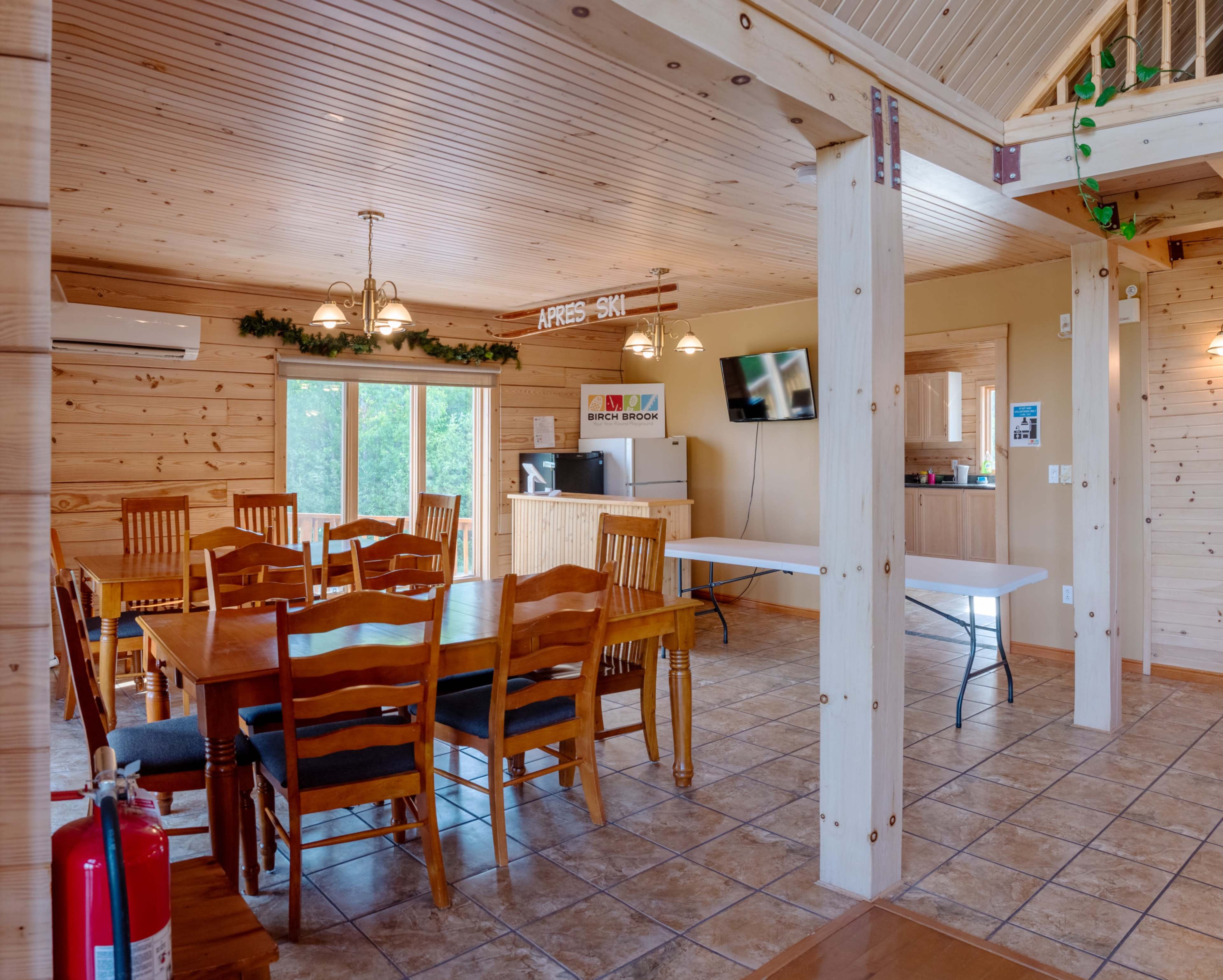 The image shows a spacious wooden interior with a dining table and chairs, a service counter, and a television mounted on the wall.