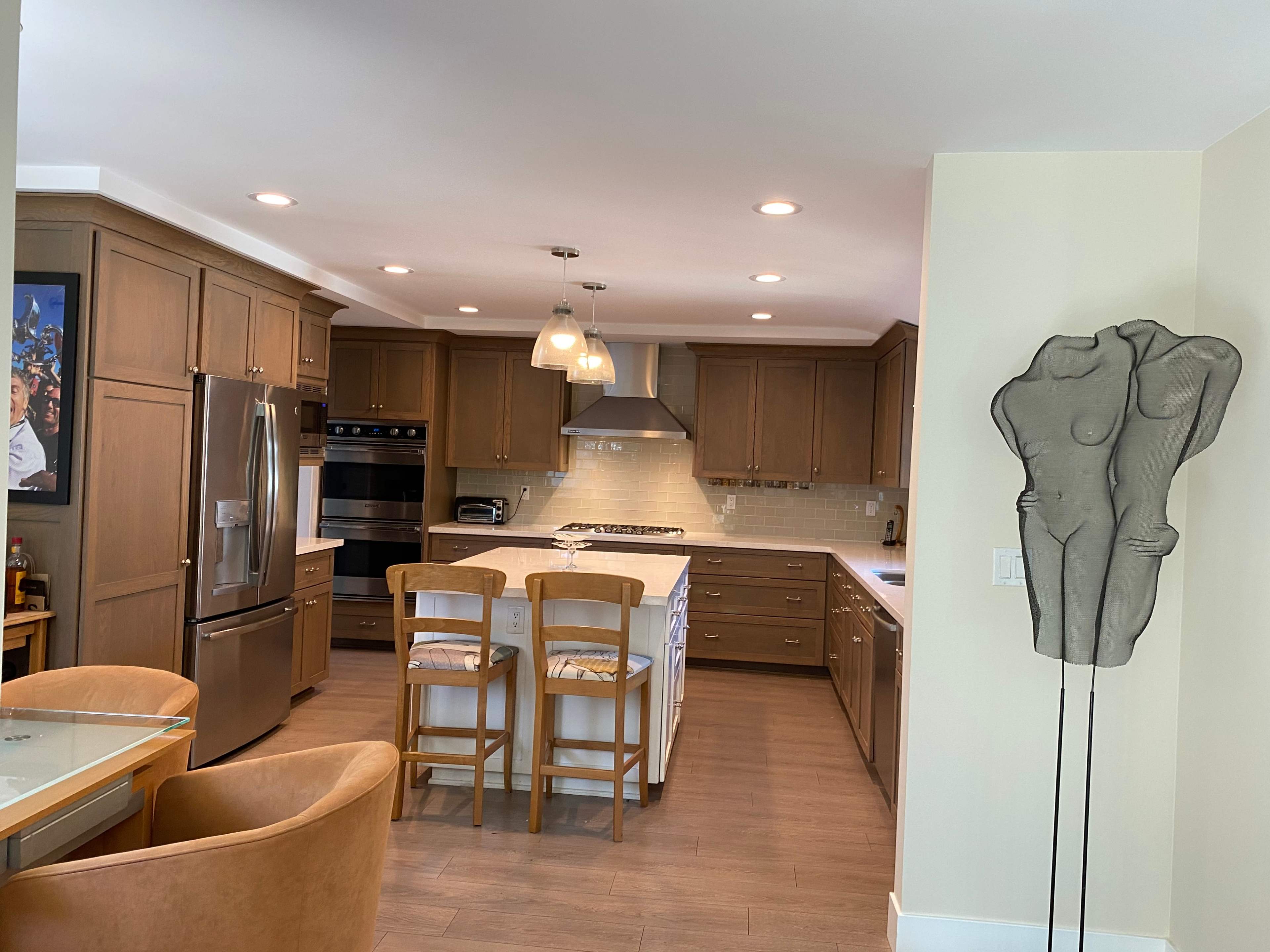 The image shows a modern kitchen with wooden cabinetry, a central island with seating, stainless steel appliances, and a decorative sculpture of a human figure nearby.