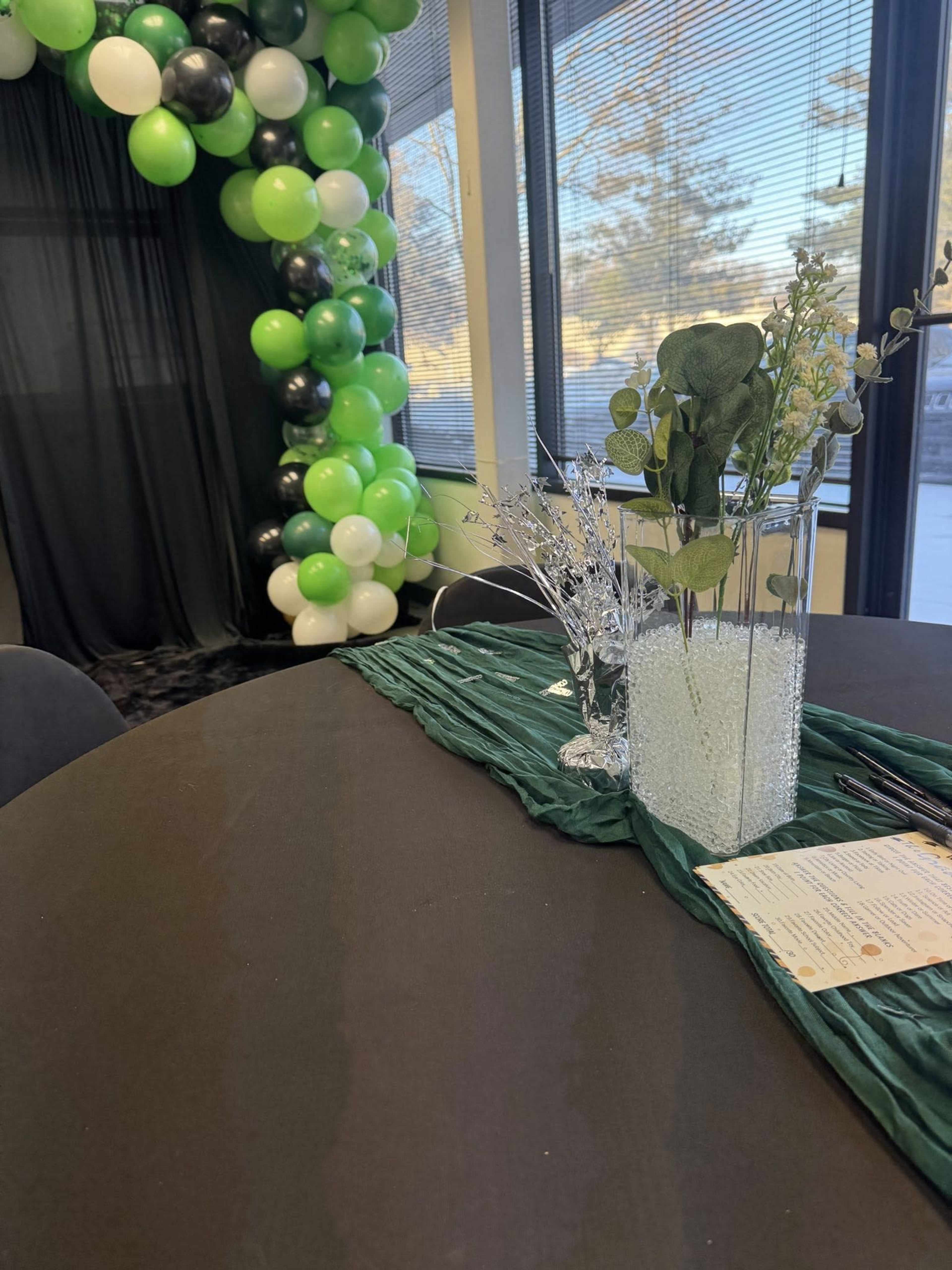 A round table is set with a green tablecloth, a floral arrangement in a clear vase, and a decorative card beside a pen, with a backdrop of black and green balloons visible in the background.
