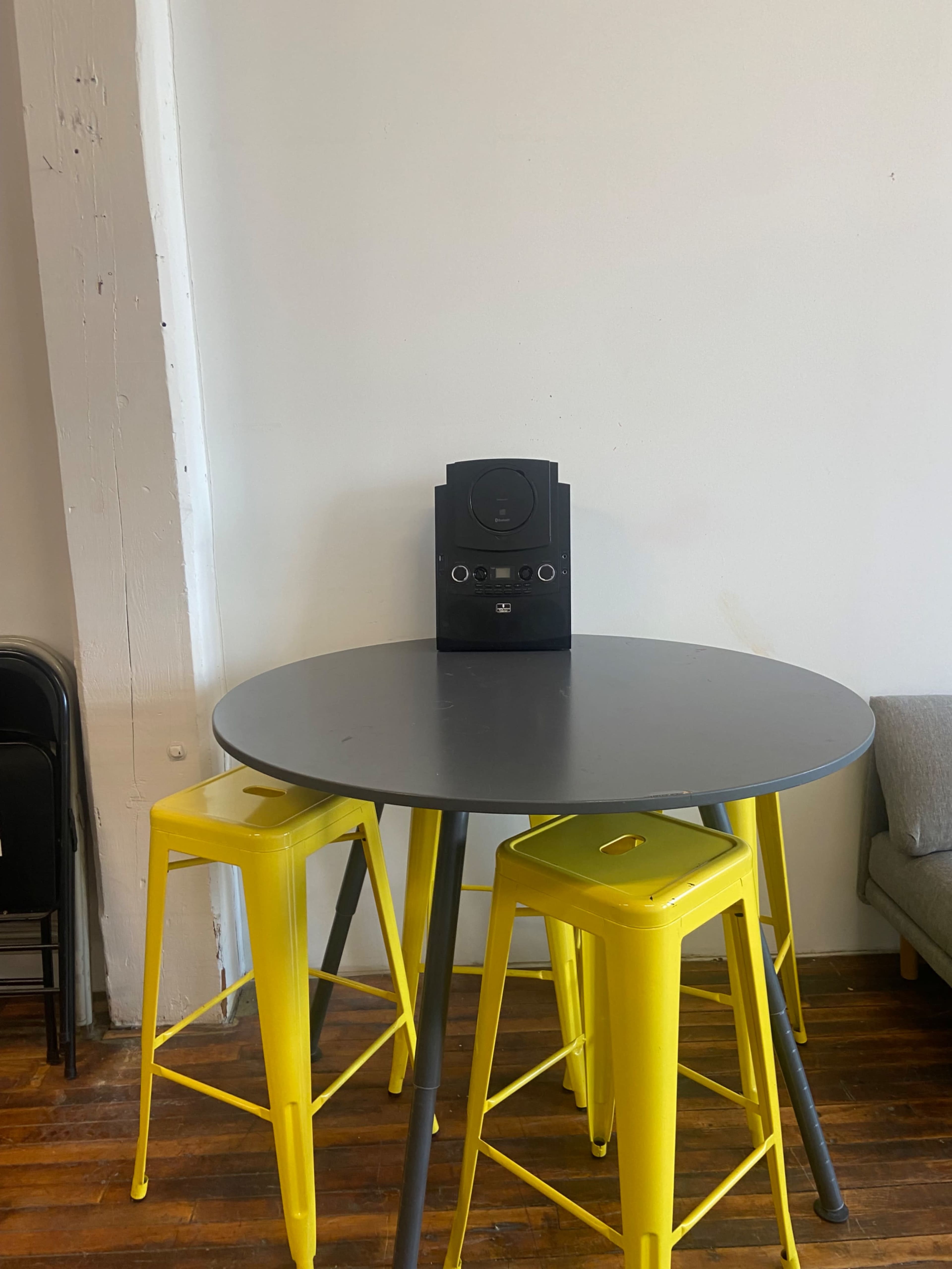 A round gray table with three yellow stools and a black speaker is positioned against a white wall.