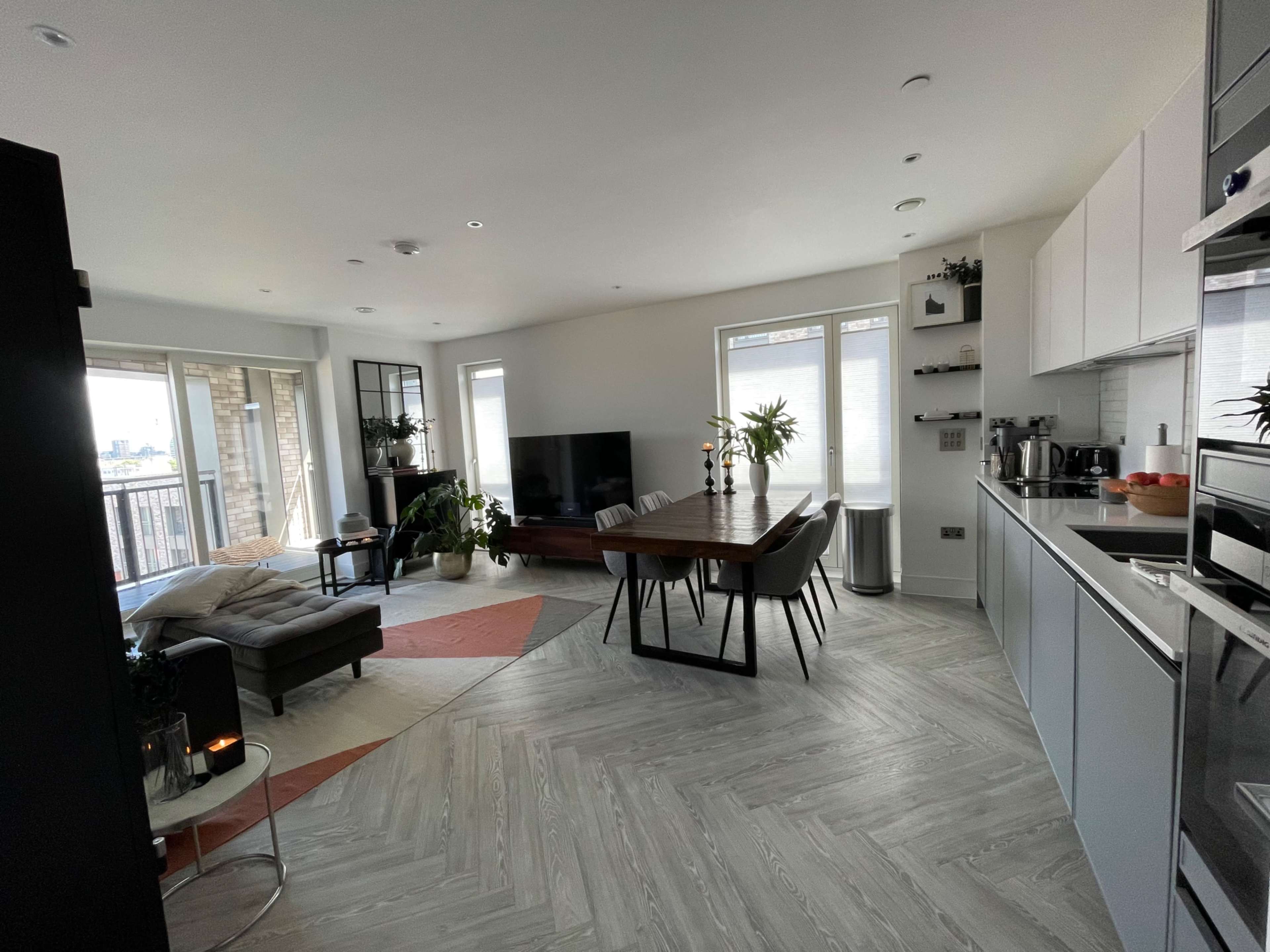 The image shows a modern open-concept kitchen and living area with a dining table, seating, and large windows allowing natural light.