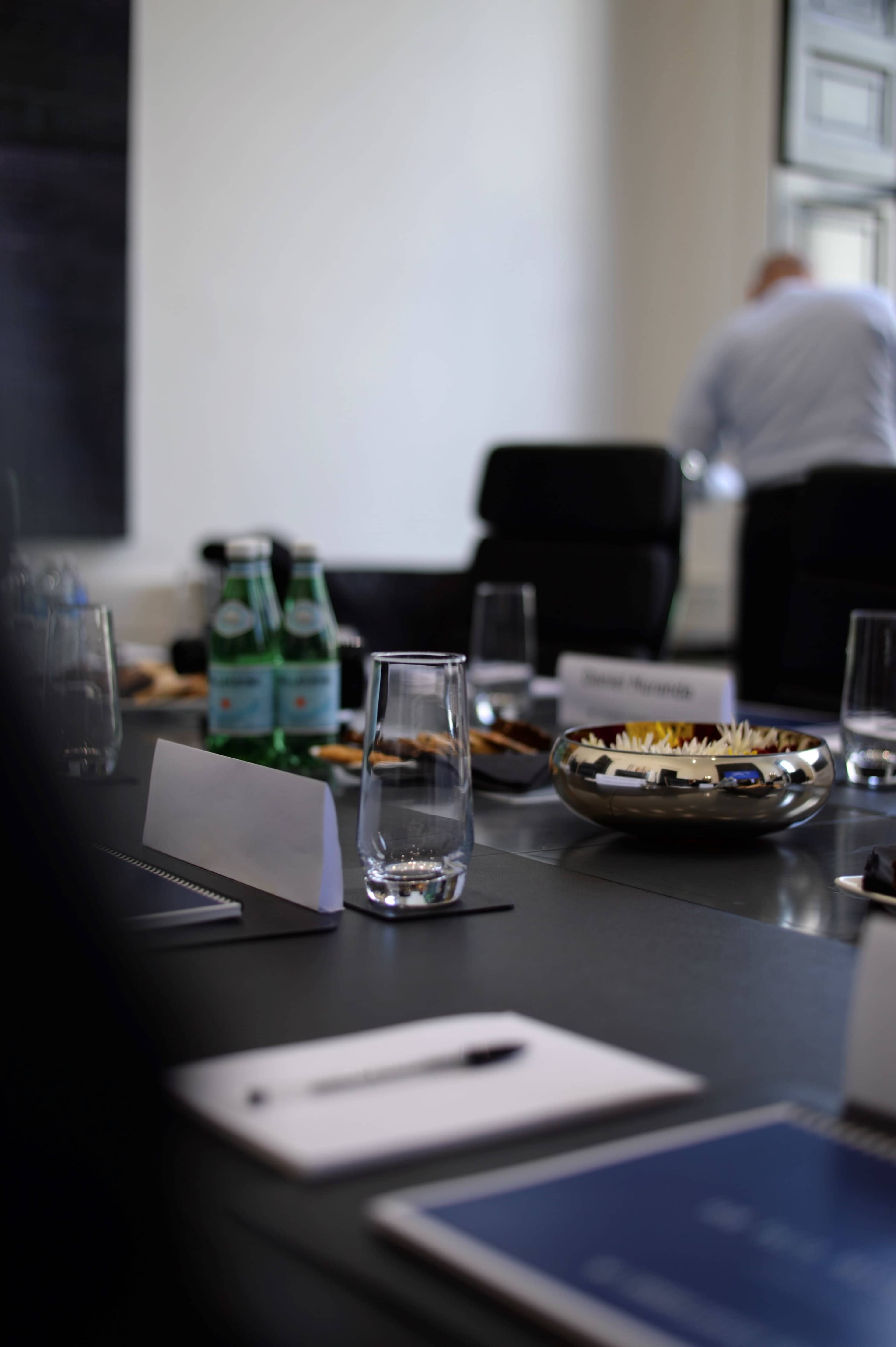 A conference room table is set with glassware, snacks, and bottles of water, while a person stands in the background.