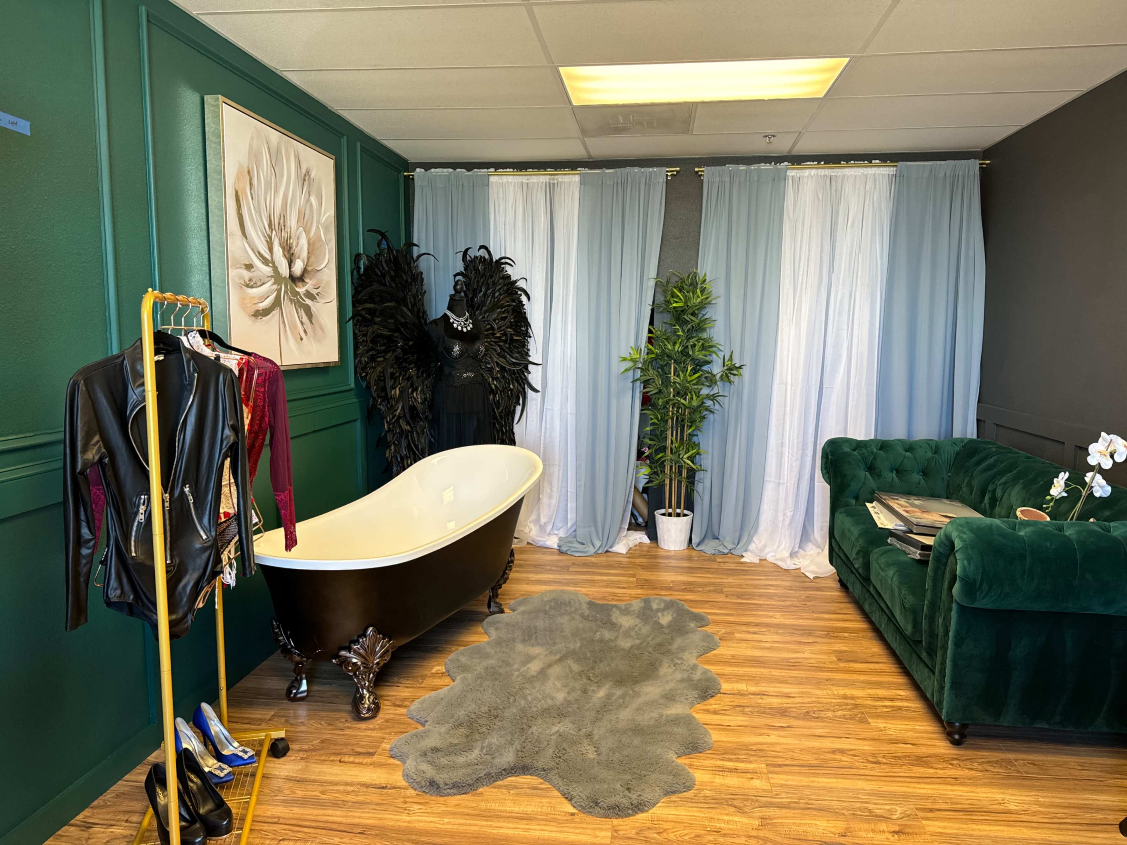 The image shows a stylish room featuring a clawfoot bathtub, a green velvet couch, a clothing rack with various outfits, a potted plant, and soft drapes dividing the space.
