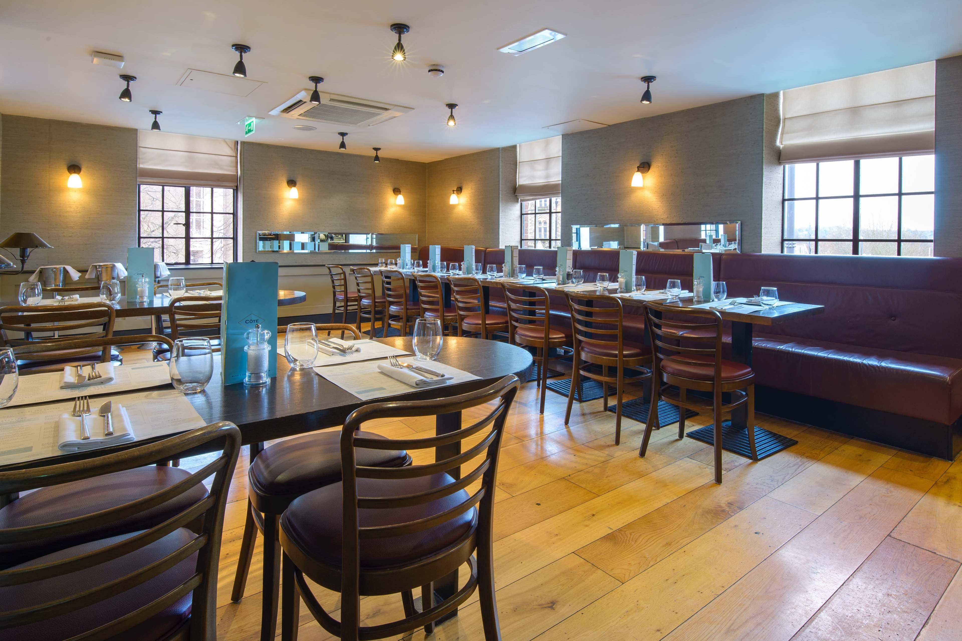 The image shows a neatly arranged restaurant interior with wooden floors, tables set with glassware and utensils, and a mix of seating options along the walls.