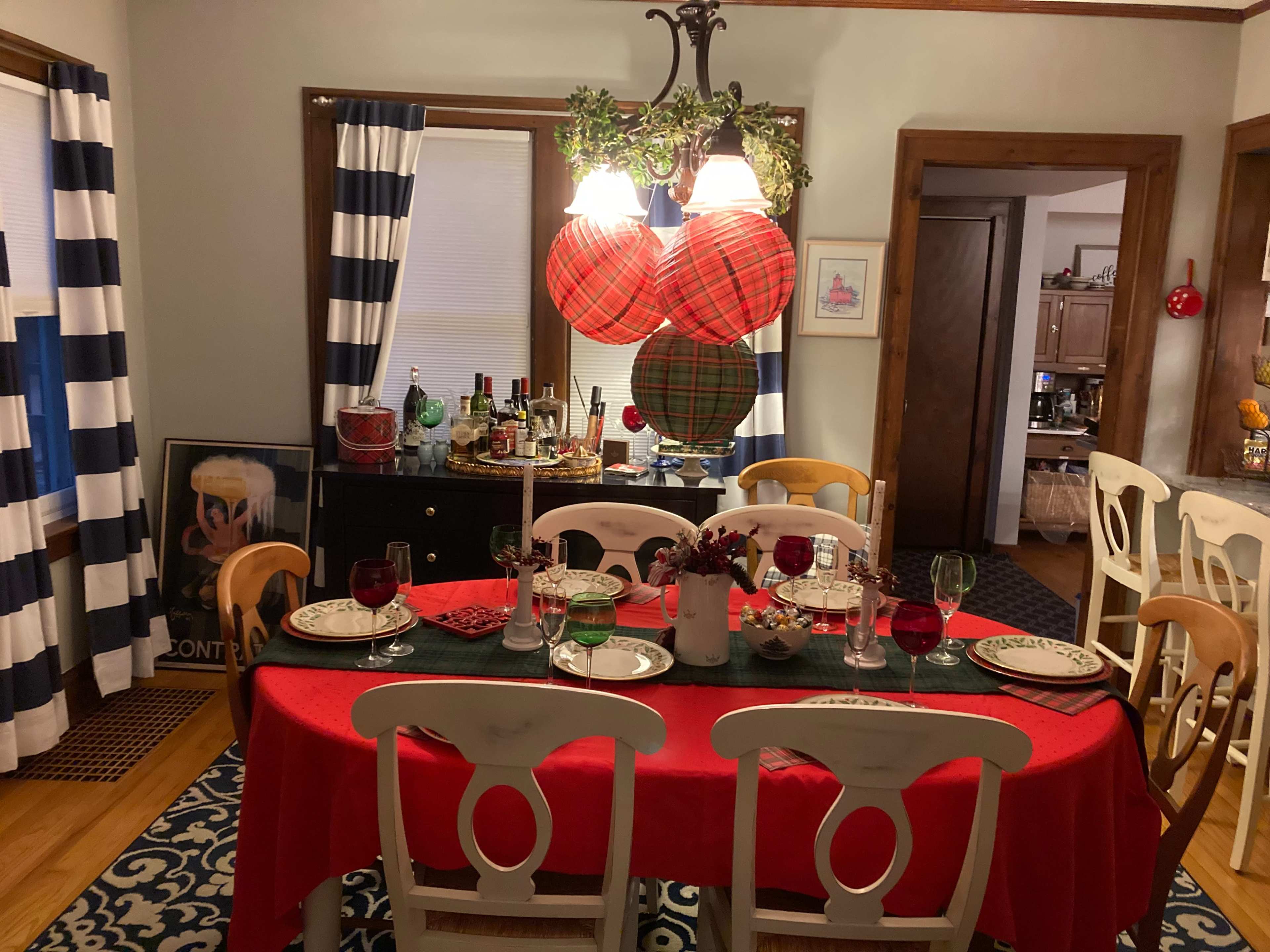 A dining room features a table set with red and green decorations, surrounded by white chairs, and adorned with a chandelier and checkered wall hangings.