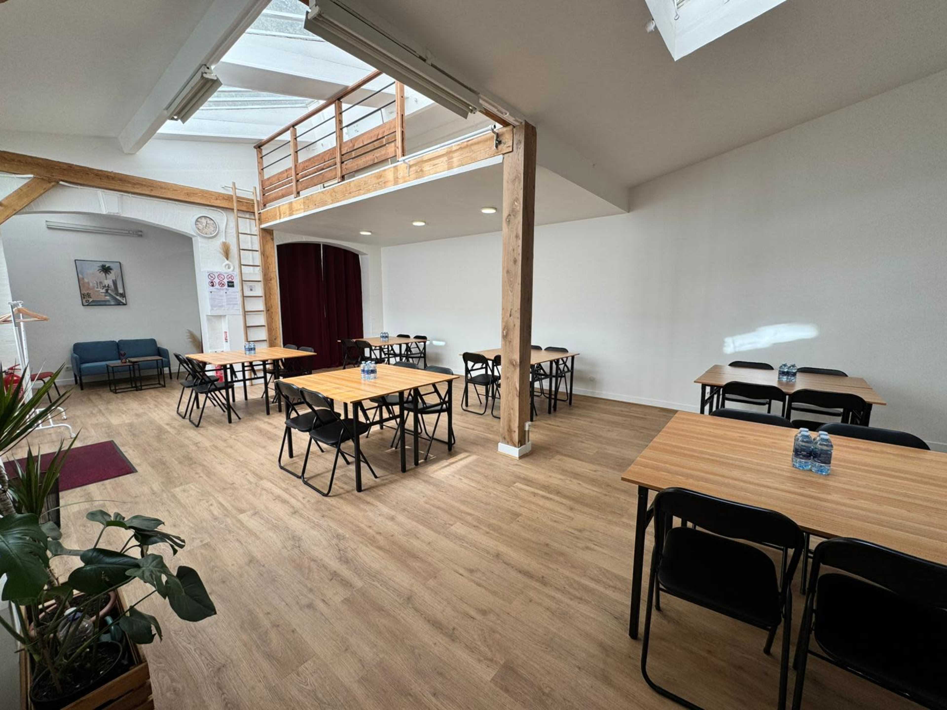The image shows a spacious, well-lit room with several wooden tables and chairs arranged in a clean layout, featuring large windows and a staircase in the background.