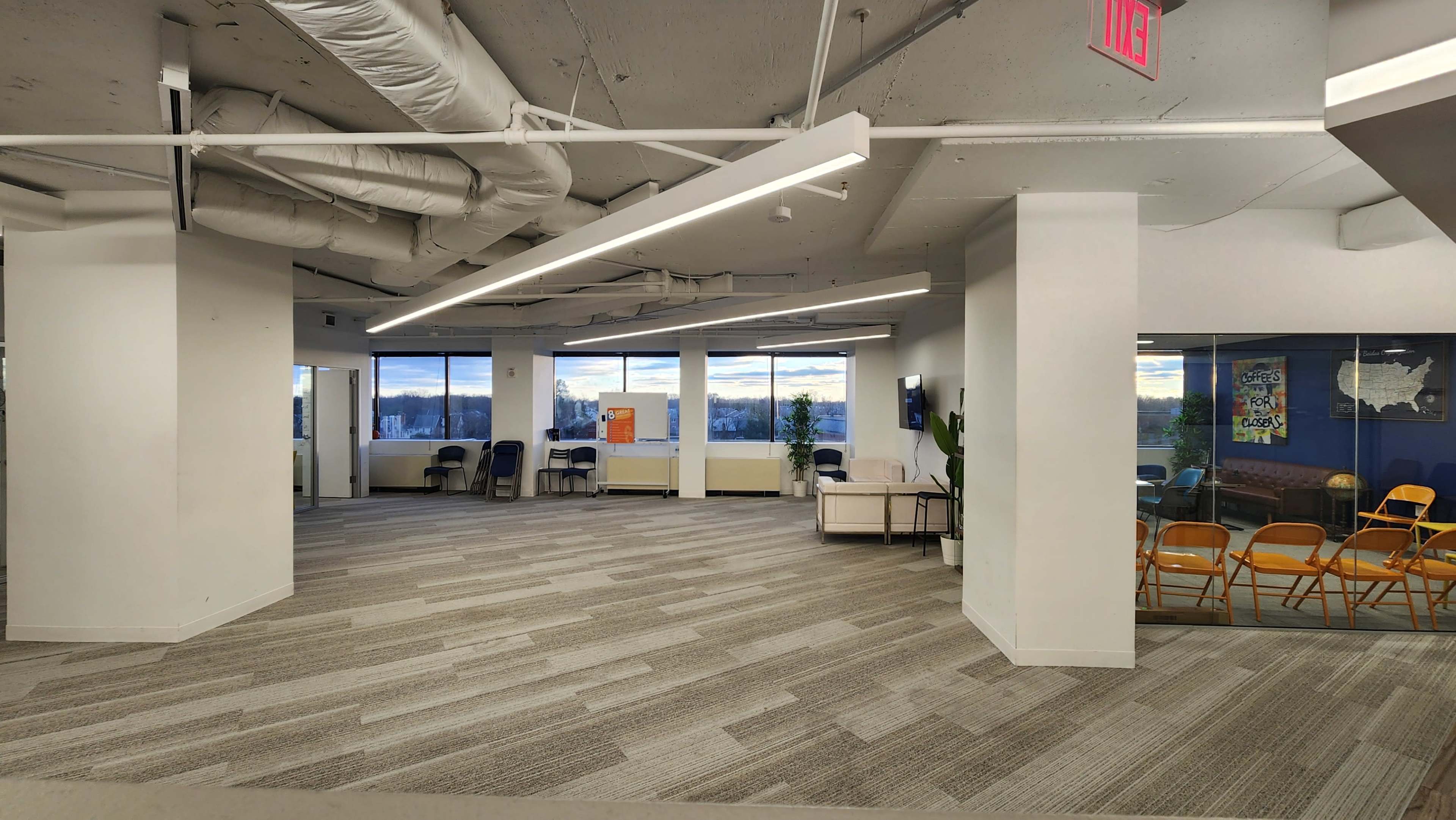 An office interior featuring several empty chairs arranged in a waiting area, large windows with a view, and a carpeted floor.