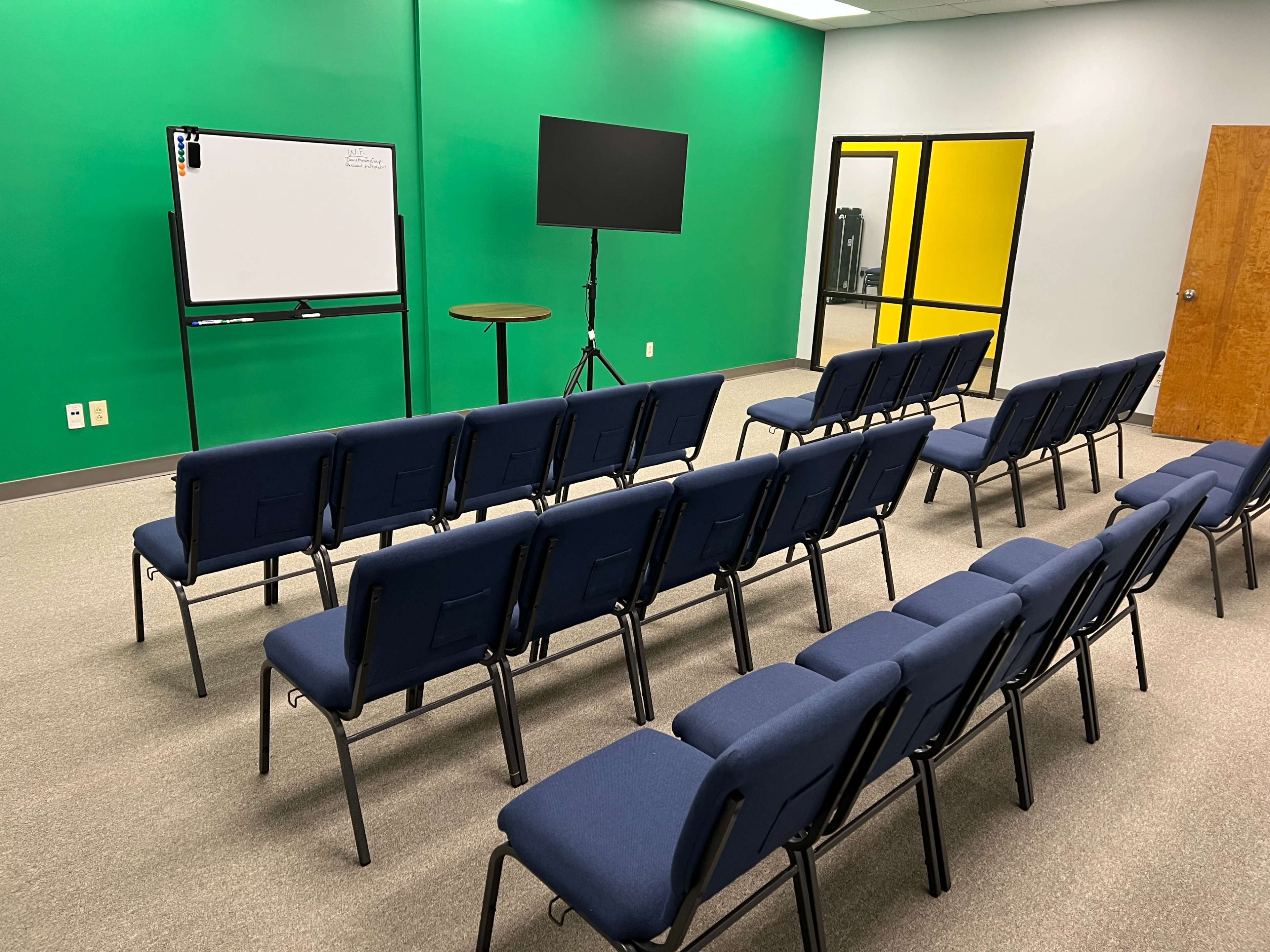 The image shows a conference room with rows of blue chairs arranged facing a whiteboard and a television on a stand against a green wall.